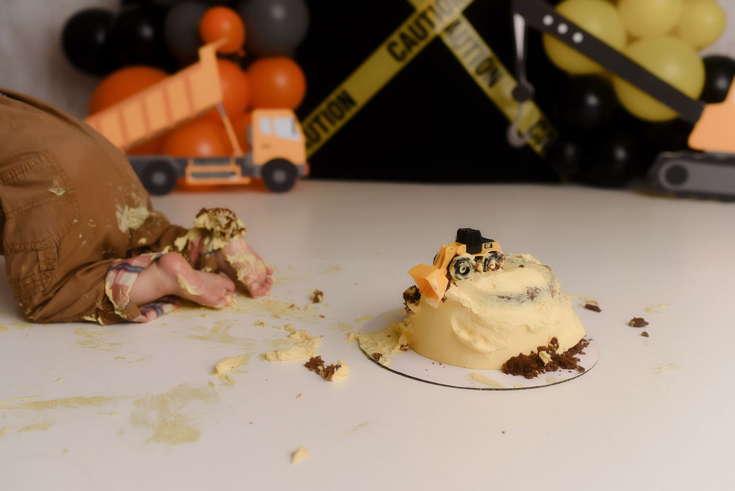 Construction-themed first birthday cake smash with a partially destroyed yellow cake, caution tape, and toy trucks in the background.