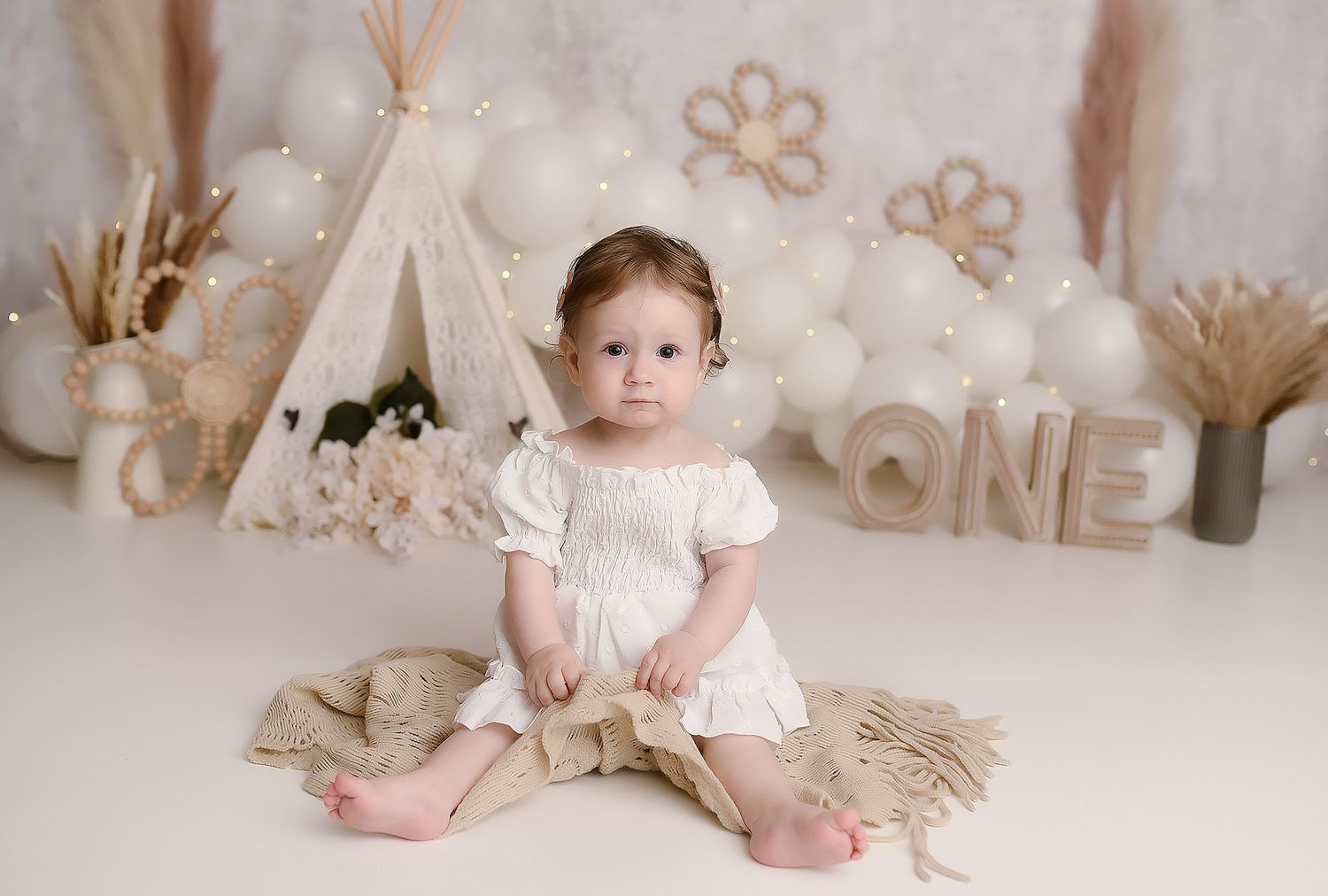 Baby girl in white dress sitting by boho teepee with ONE sign and white balloon backdrop.