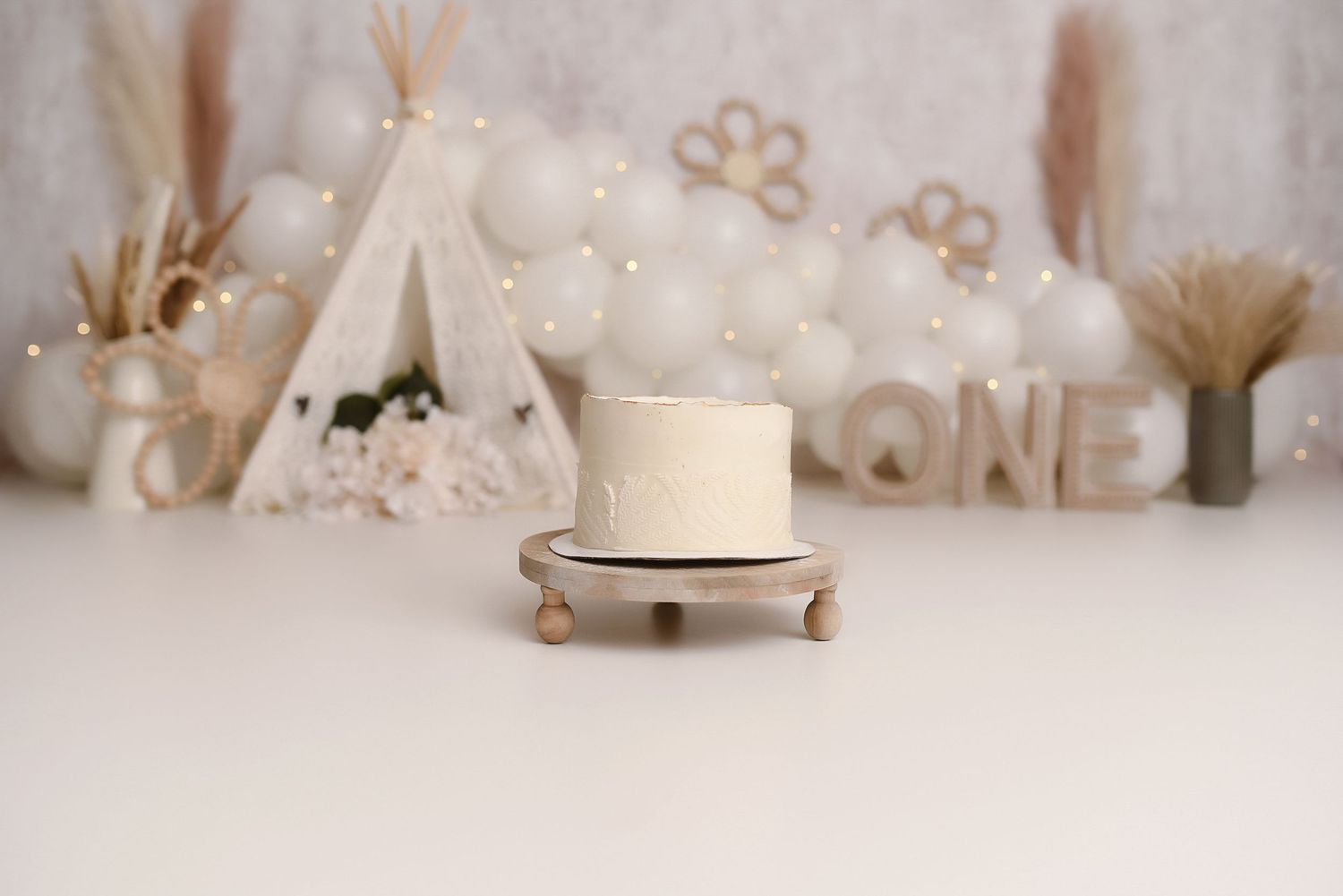 Elegant white first birthday smash cake on wooden stand with boho teepee and balloon arch backdrop.