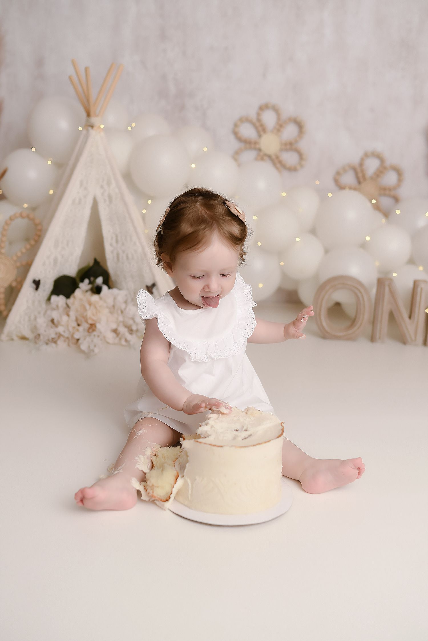 One year old girl in white dress smashing cream cake at boho teepee first birthday session.