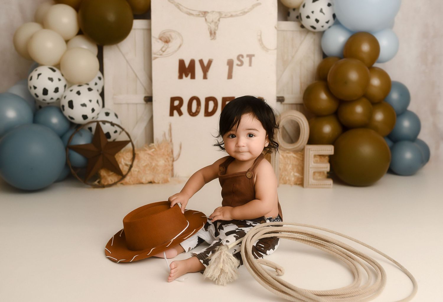 Baby in brown romper sits at My 1st Rodeo themed photo shoot with balloons, cowboy hat, and lasso.