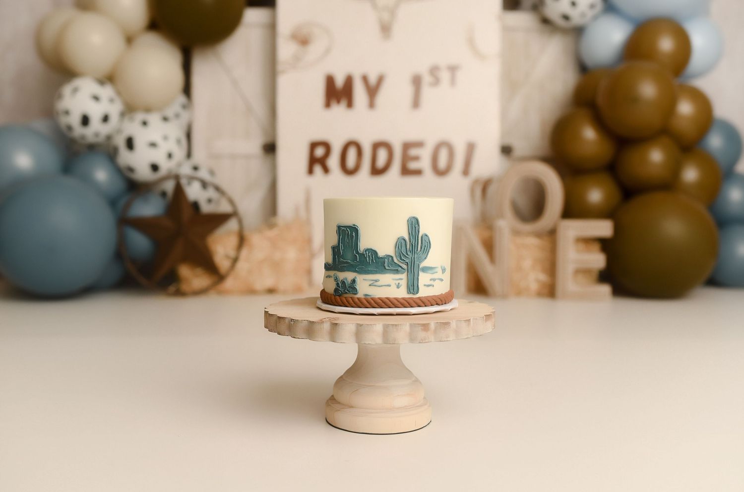 A first birthday smash cake with cactus design on a wooden stand, surrounded by balloons and a My 1st Rodeo sign.