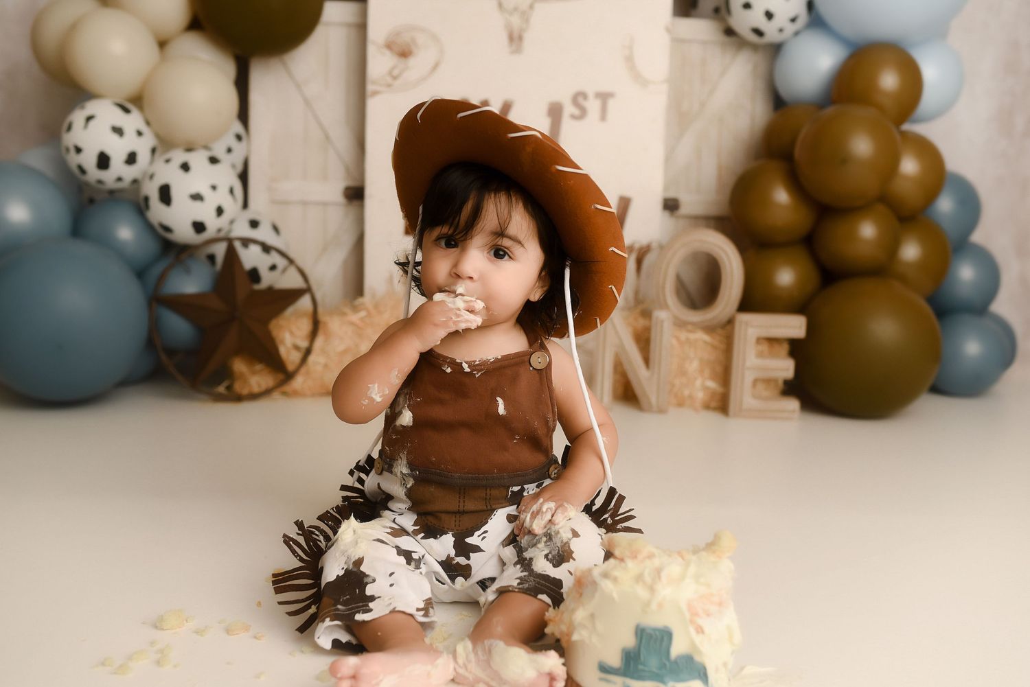 Baby in cowboy hat enjoys first birthday cake smash with western themed balloons and ONE sign backdrop.