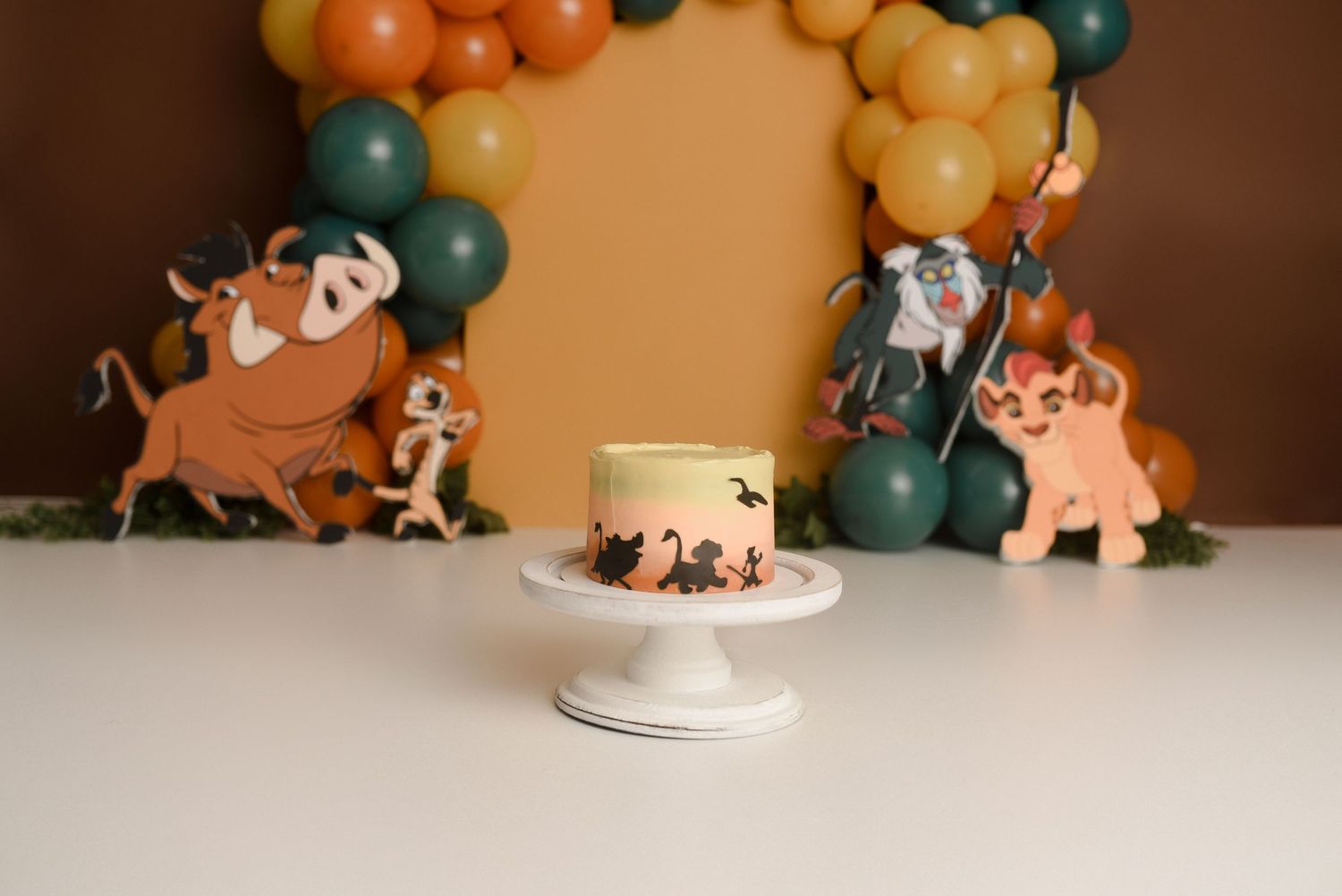 Lion King themed first birthday cake smash setup with character cutouts and colorful balloon arch backdrop.
