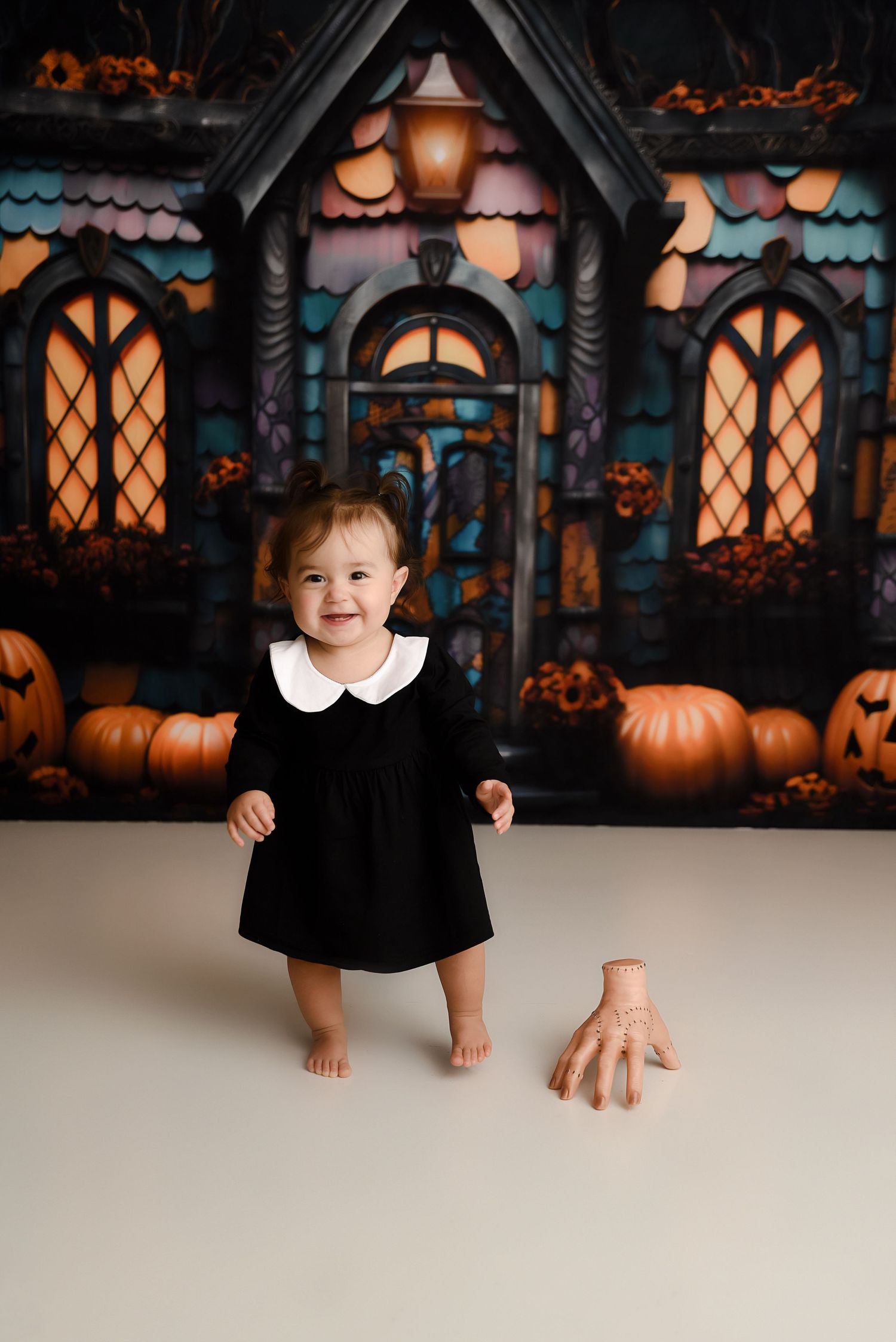 Smiling baby in black dress with white collar standing before Halloween haunted house backdrop.