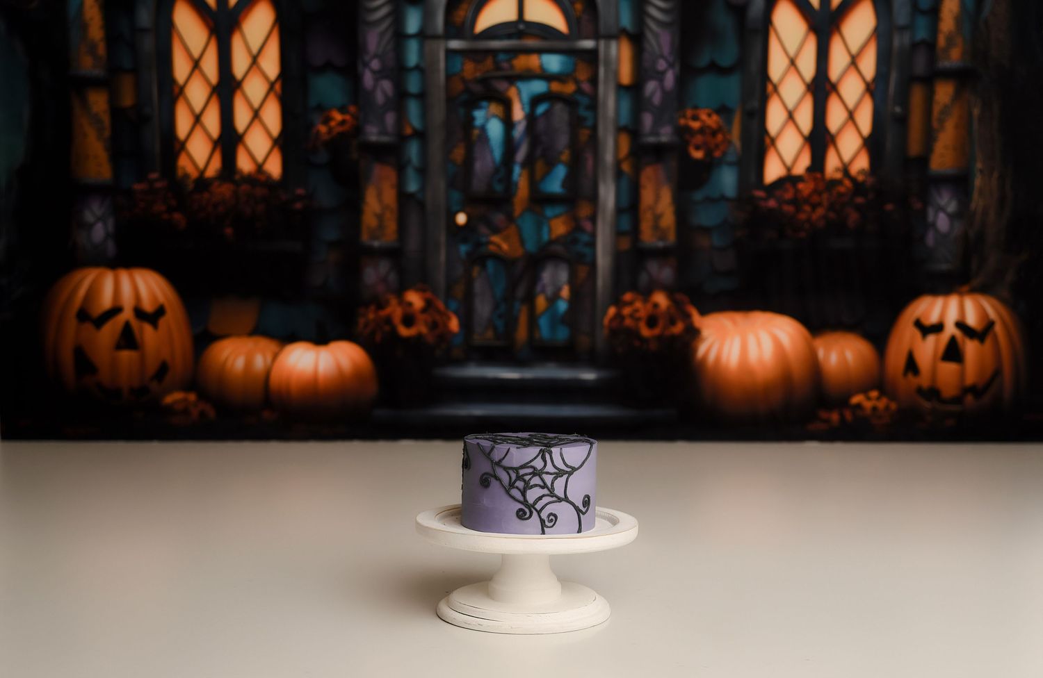 Purple spider web first birthday cake on pedestal with Halloween haunted house pumpkin backdrop.