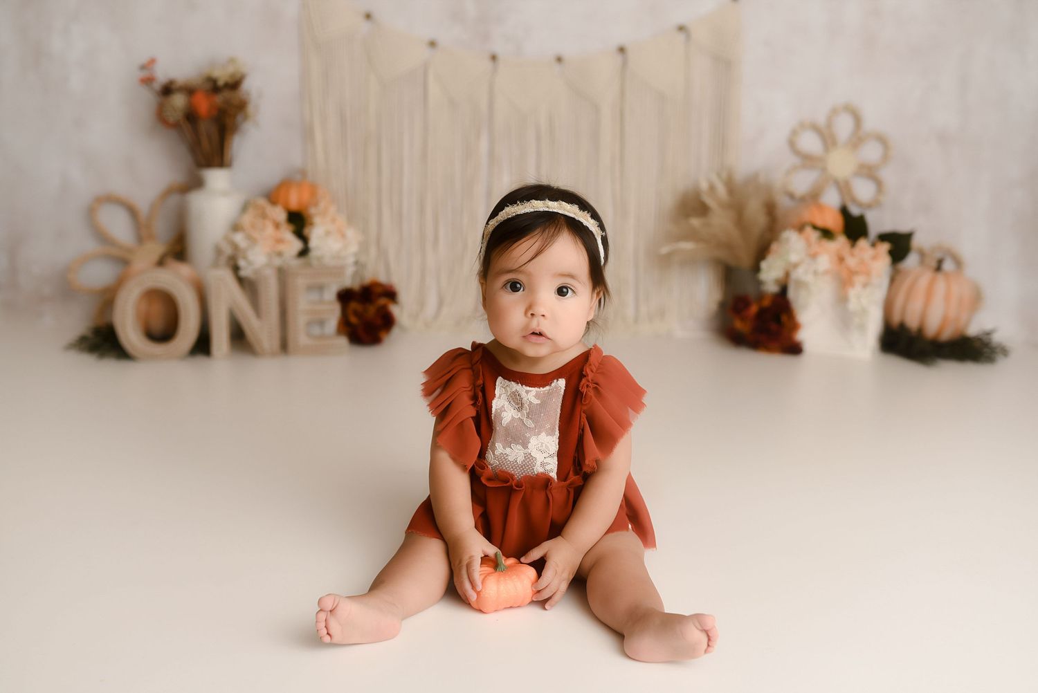 Baby girl in rust romper holding pumpkin with boho ONE birthday backdrop.
