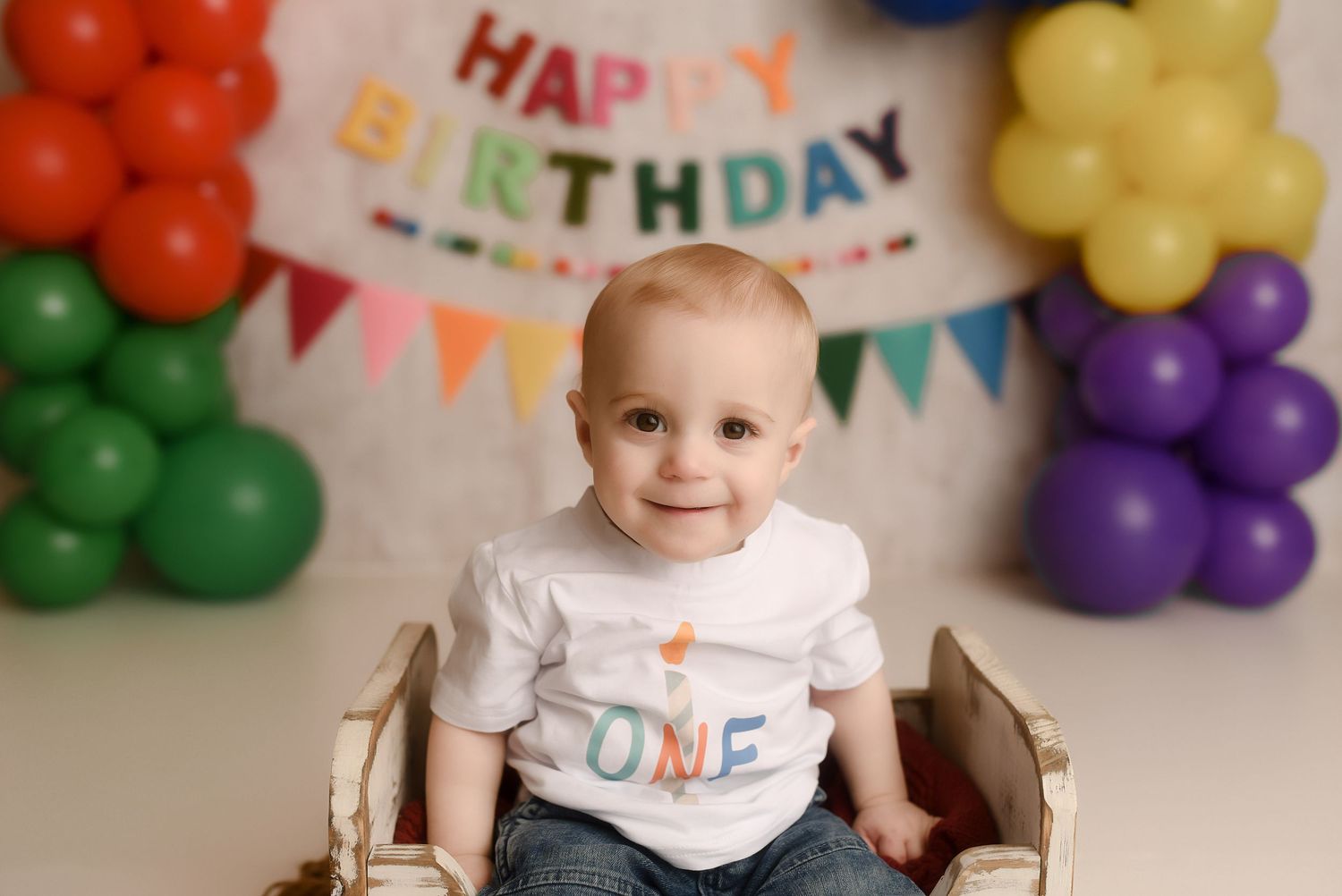 A smiling baby wearing a white ONE shirt sits in a wooden crate surrounded by colorful balloons at a first birthday party.