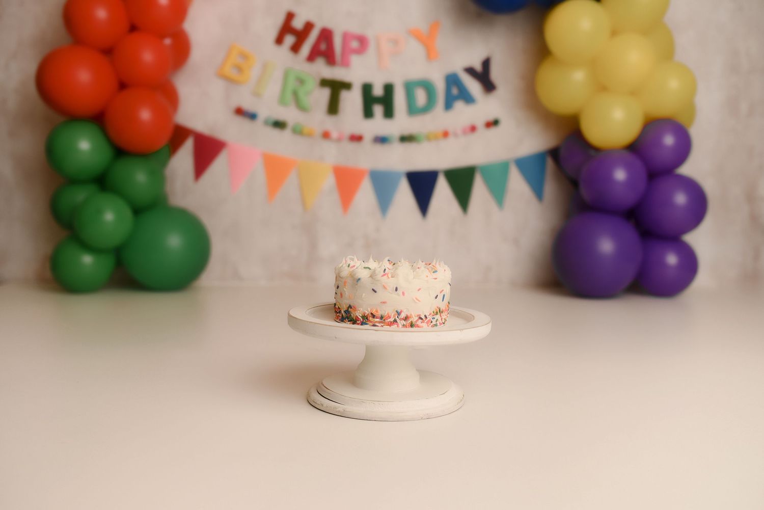 White birthday cake on pedestal stand with colorful balloon columns and Happy Birthday banner backdrop.