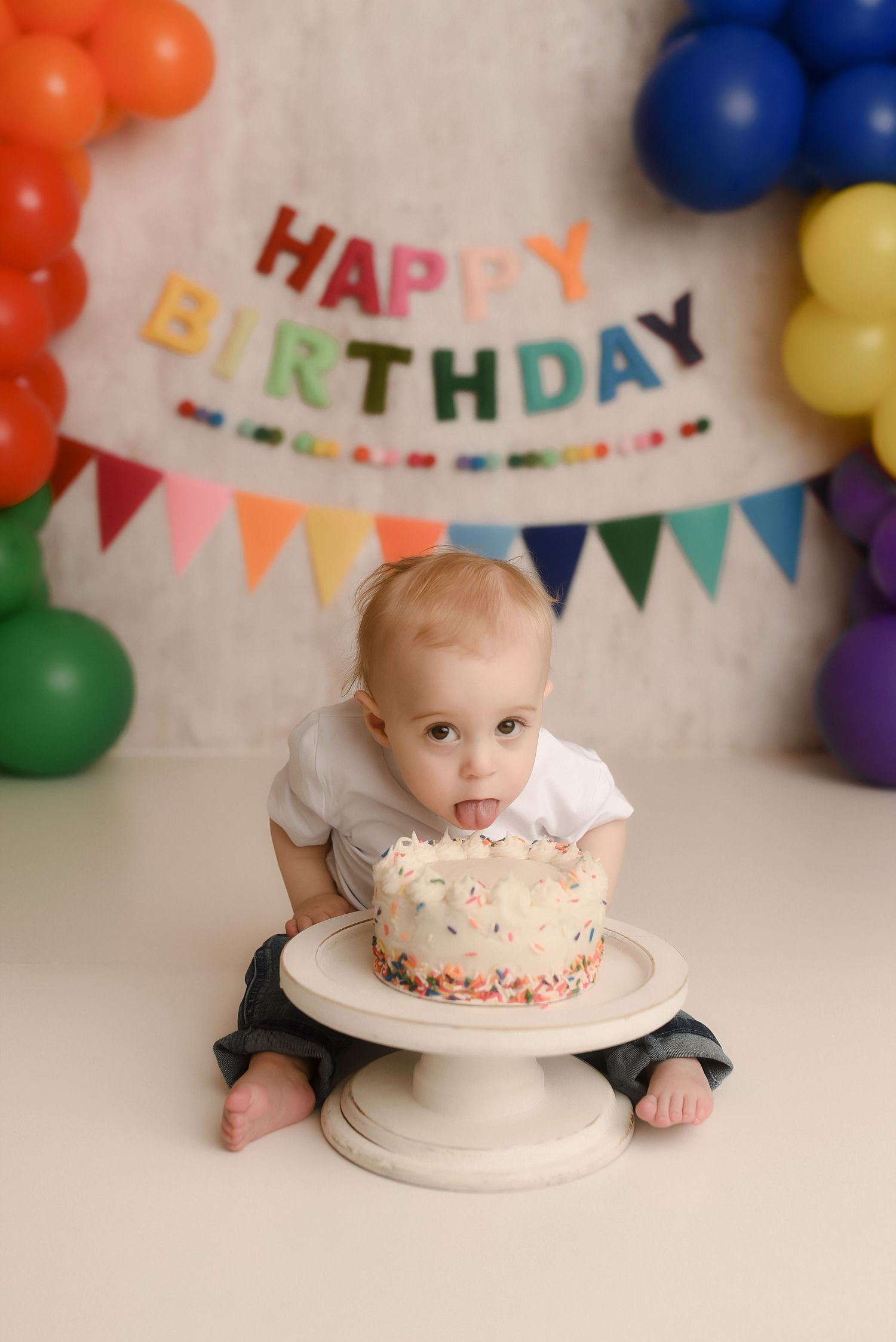 Baby's first birthday cake smash photo with colorful balloon arch and Happy Birthday banner backdrop.