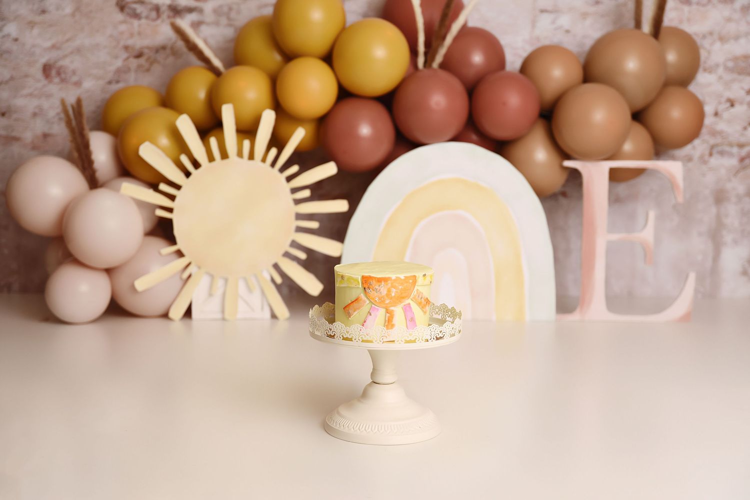 Boho sunshine first birthday smash cake with rainbow, sun decor and warm balloon garland.