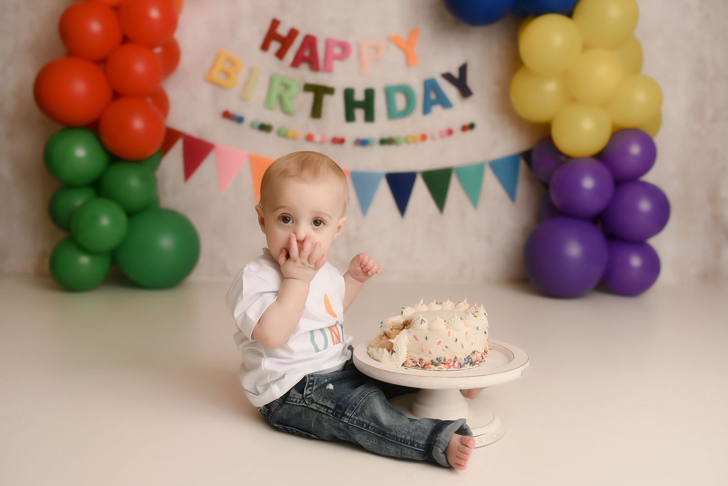 Baby's first birthday cake smash with colorful balloons and Happy Birthday banner on white backdrop.