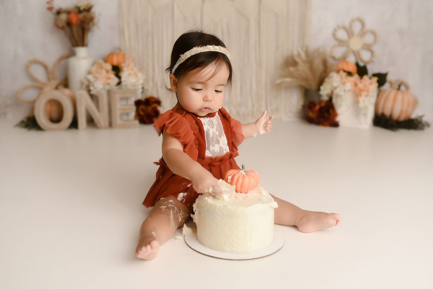 Baby girl in rust romper smashing white cake with boho ONE birthday backdrop.