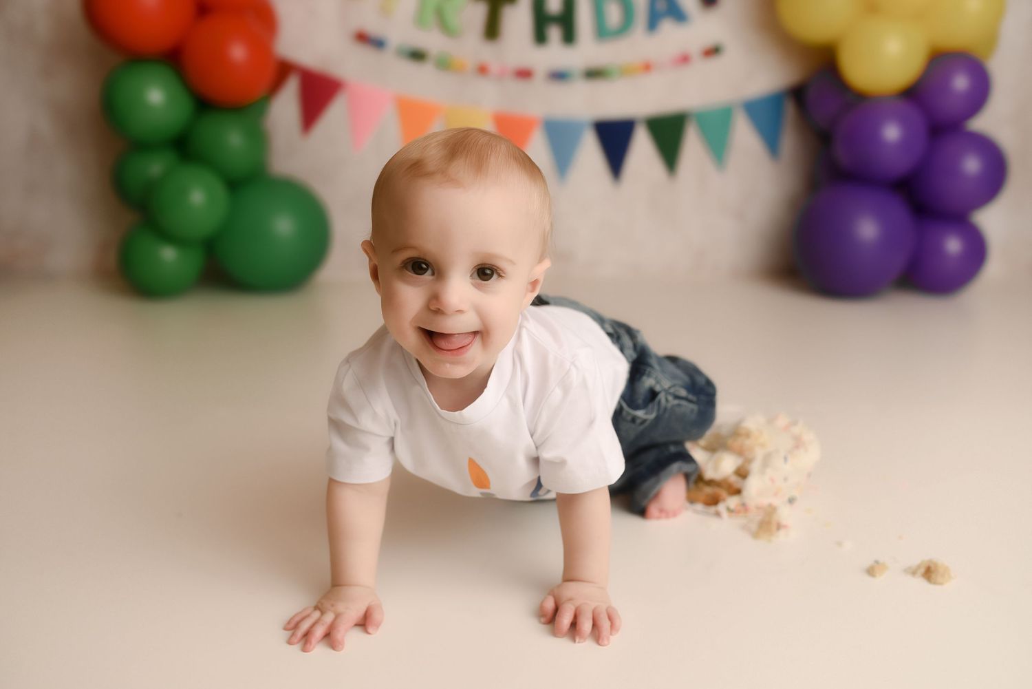 Smiling baby crawling on white floor at first birthday party with colorful balloons and bunting banner.
