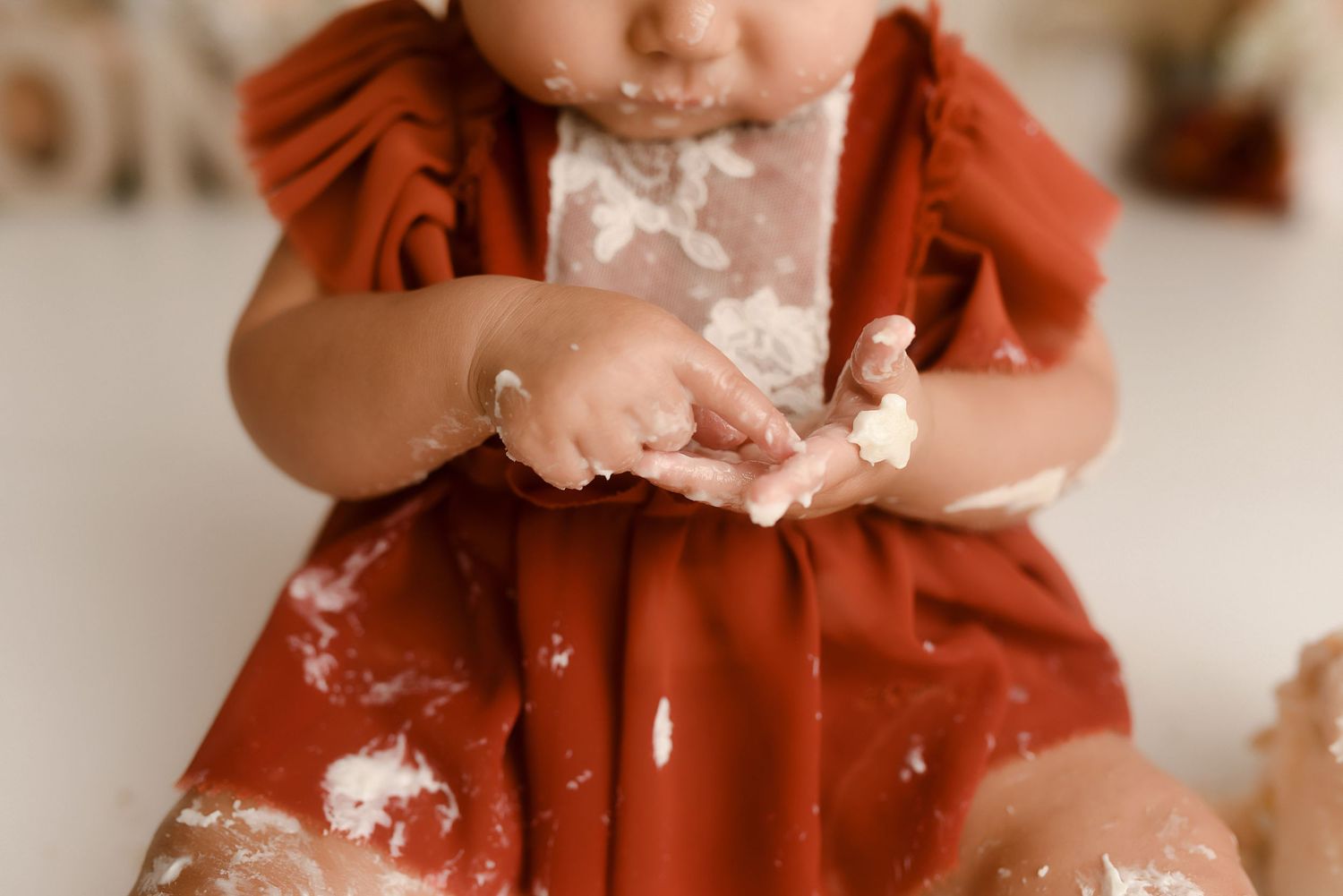 Close-up of baby girl in rust dress with cake-covered hands during smash session.