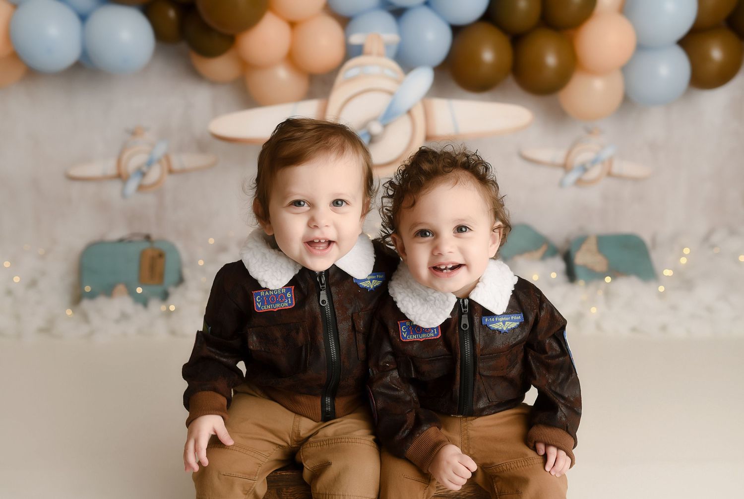 Twin babies in matching brown aviator jackets smiling at cake smash session with airplane balloon backdrop.