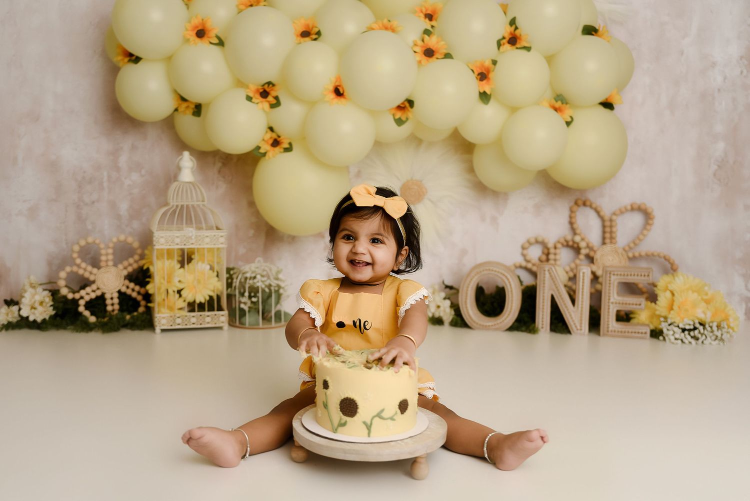 Smiling baby girl in yellow outfit celebrates first birthday with sunflower cake smash and balloon backdrop.