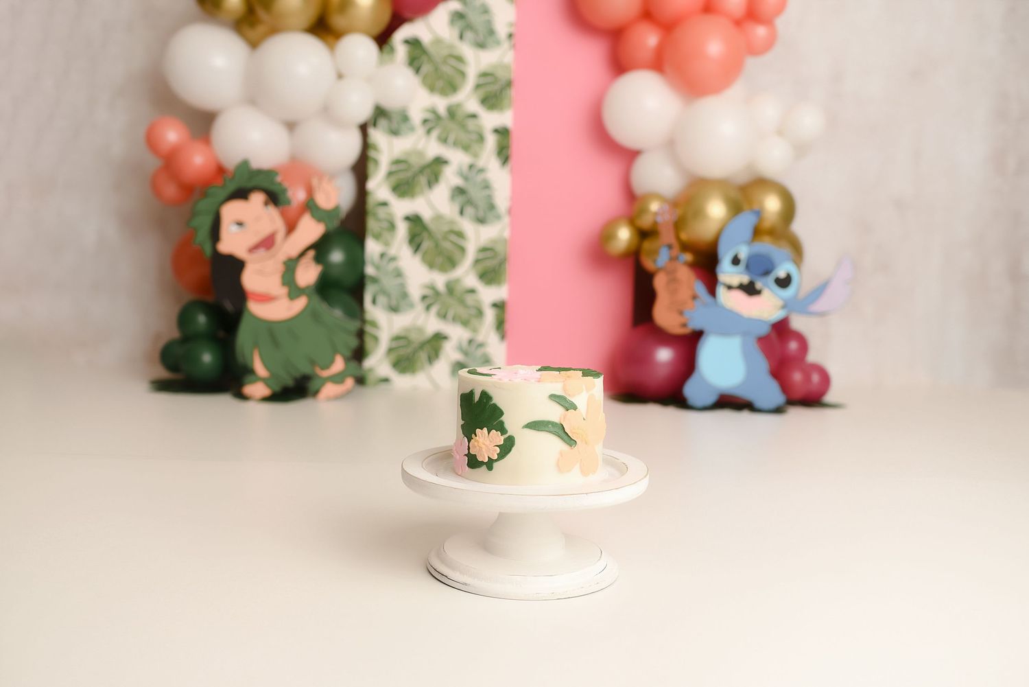 Lilo and Stitch first birthday cake smash setup with tropical cake on white stand and character decorations.