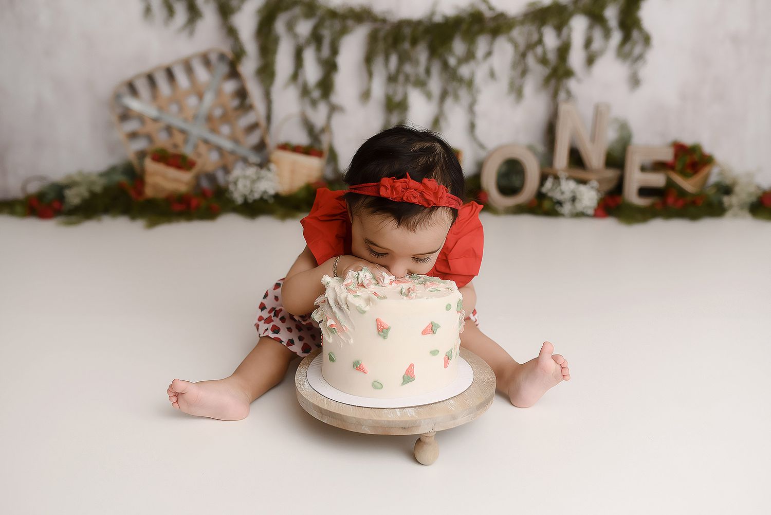 Baby girl in red outfit leaning into white cake at rustic ONE birthday cake smash session.