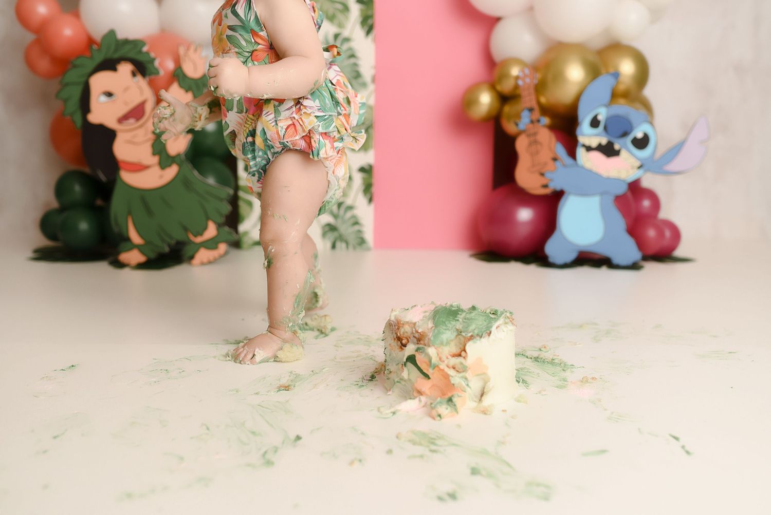 Baby's feet standing in smashed Lilo and Stitch themed birthday cake on white floor with balloon backdrop.