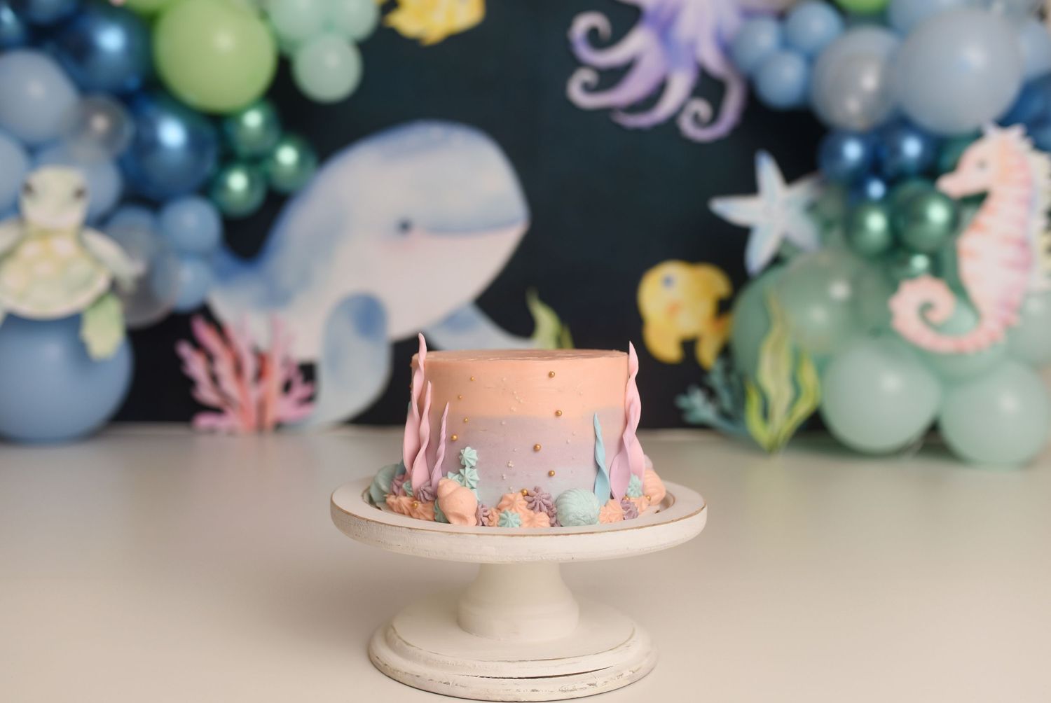 Pink ocean-themed first birthday smash cake with sea creature decorations on white stand and balloon backdrop.