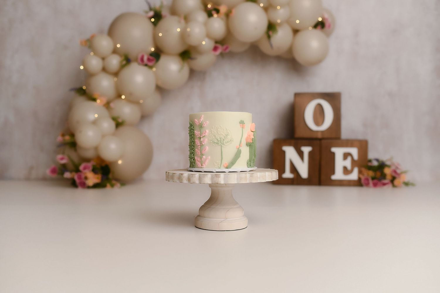 First birthday smash cake on white stand with ONE blocks and balloon arch backdrop.