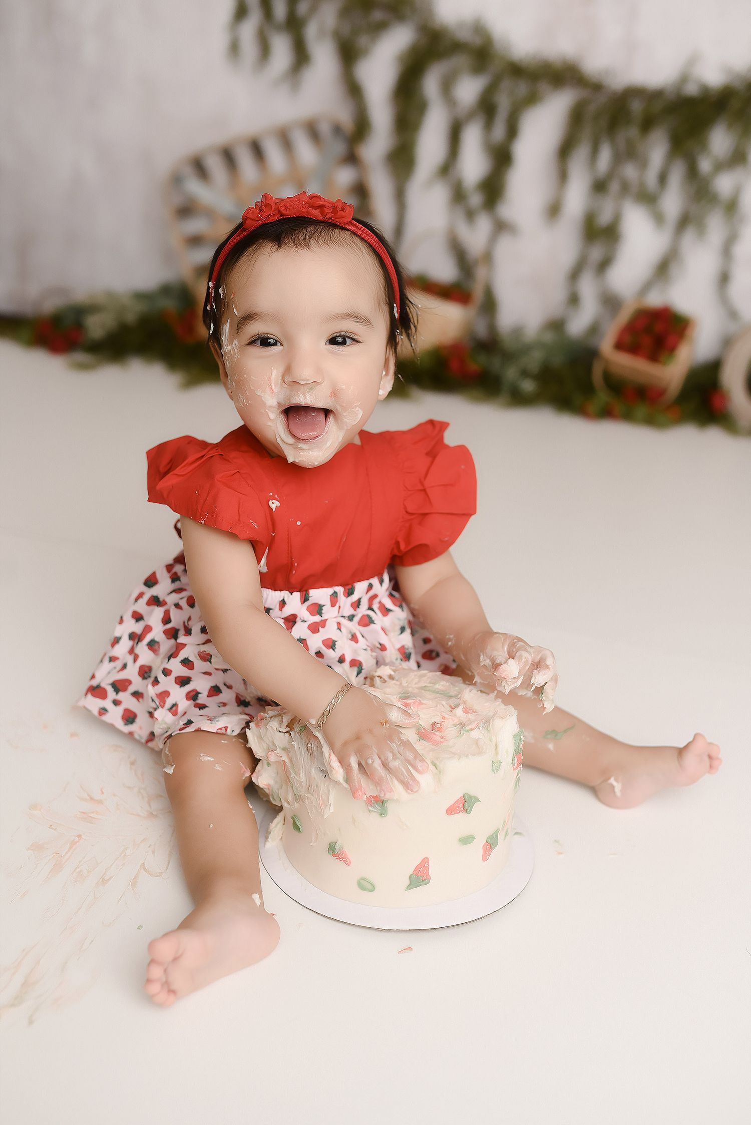 Happy baby girl laughing with cake on hands during strawberry themed first birthday cake smash.