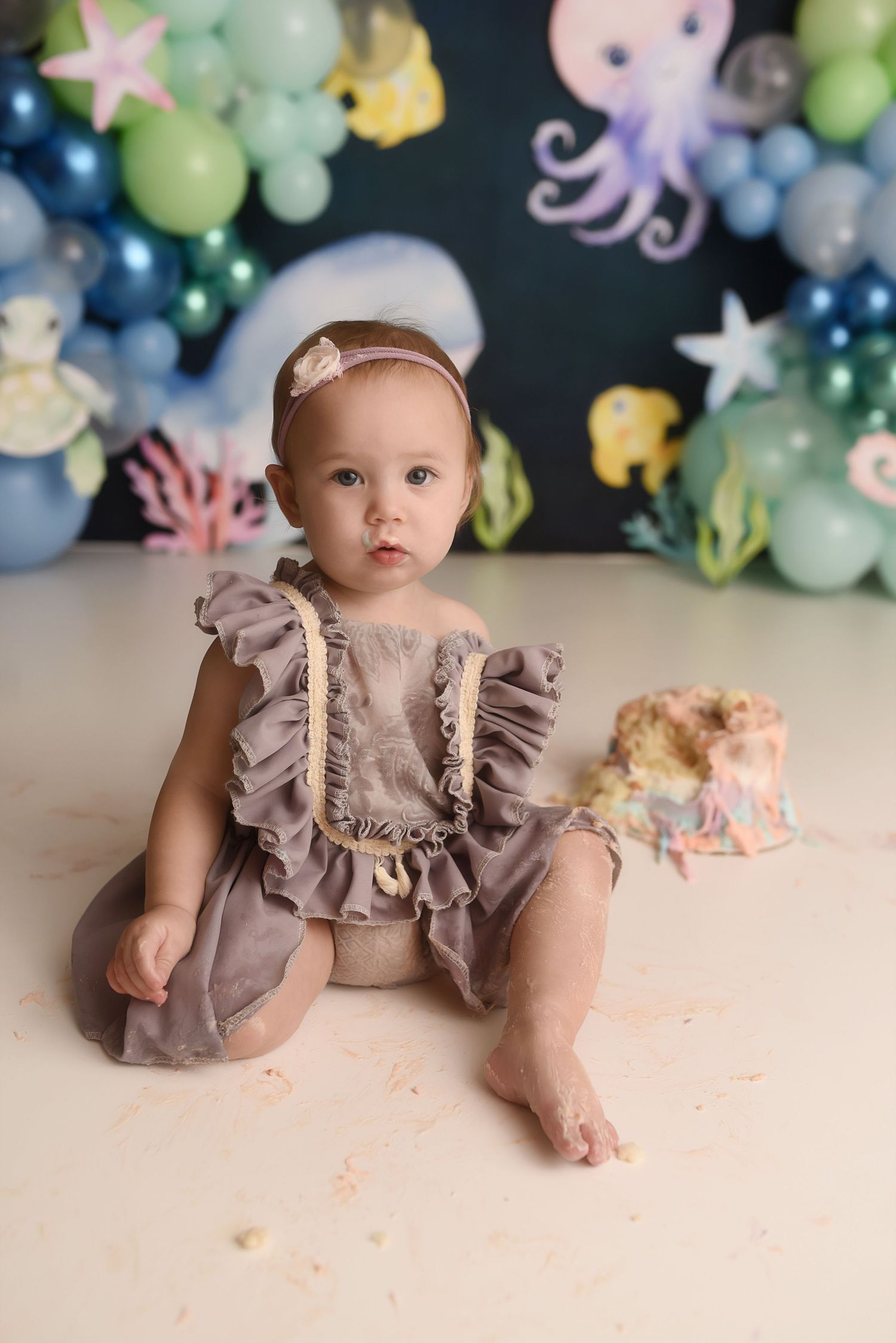 Baby girl in gray ruffle romper sitting beside smashed ocean-themed birthday cake with balloon backdrop.