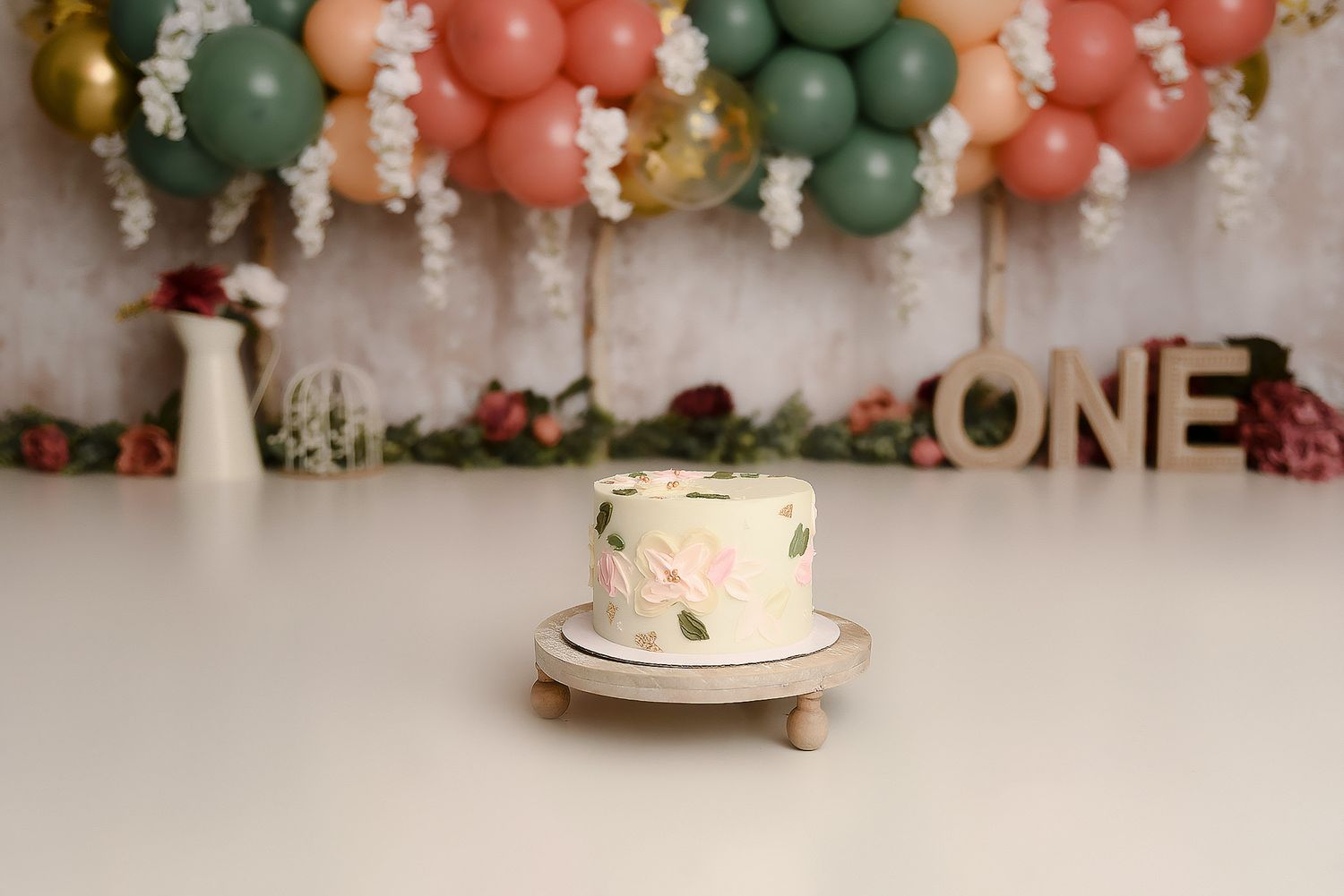 Elegant floral first birthday cake on wood stand with peach green gold balloon garland and ONE sign.