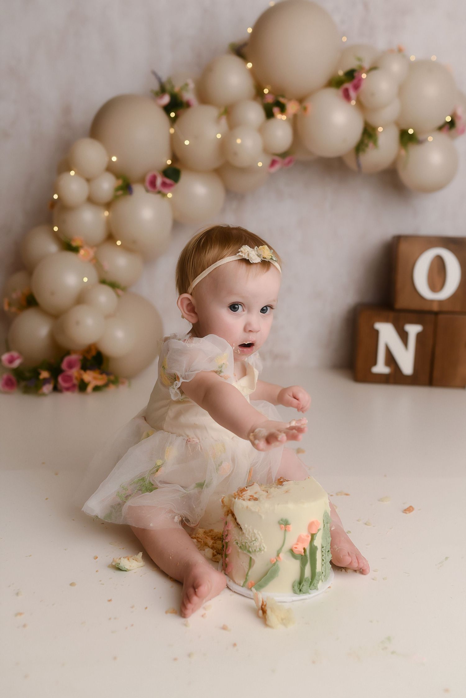 Baby girl in white dress smashes first birthday cake surrounded by beige balloons and floral decorations spelling ONE.