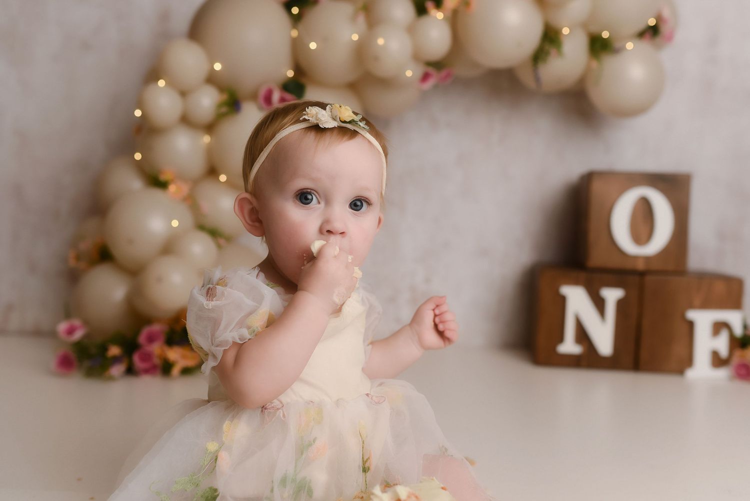 Baby girl in a white tulle dress sits before balloon arch and ONE blocks at her first birthday cake smash photo session.