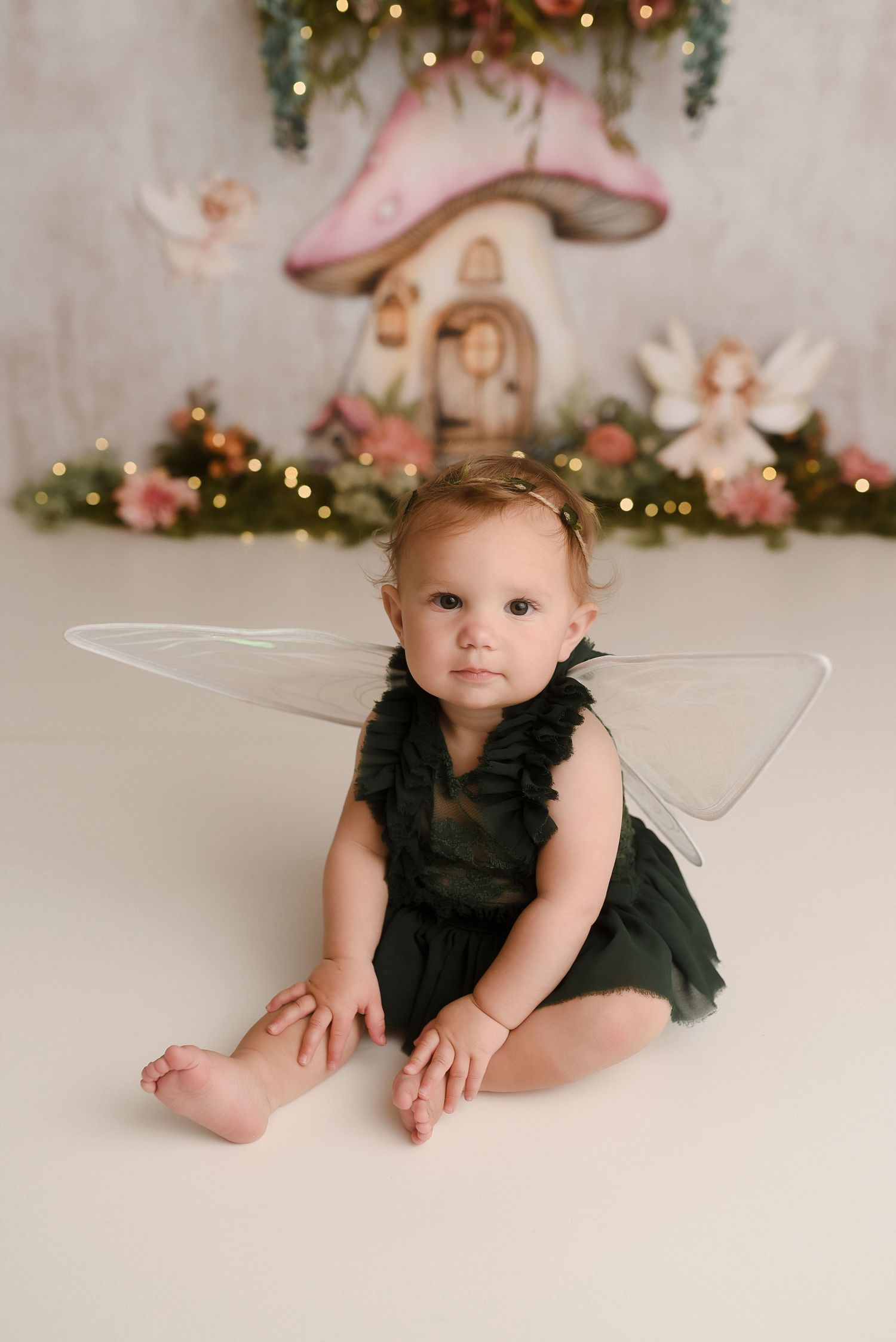 Baby in dark green fairy costume with sheer wings sits before enchanting mushroom house backdrop with soft bokeh lights.