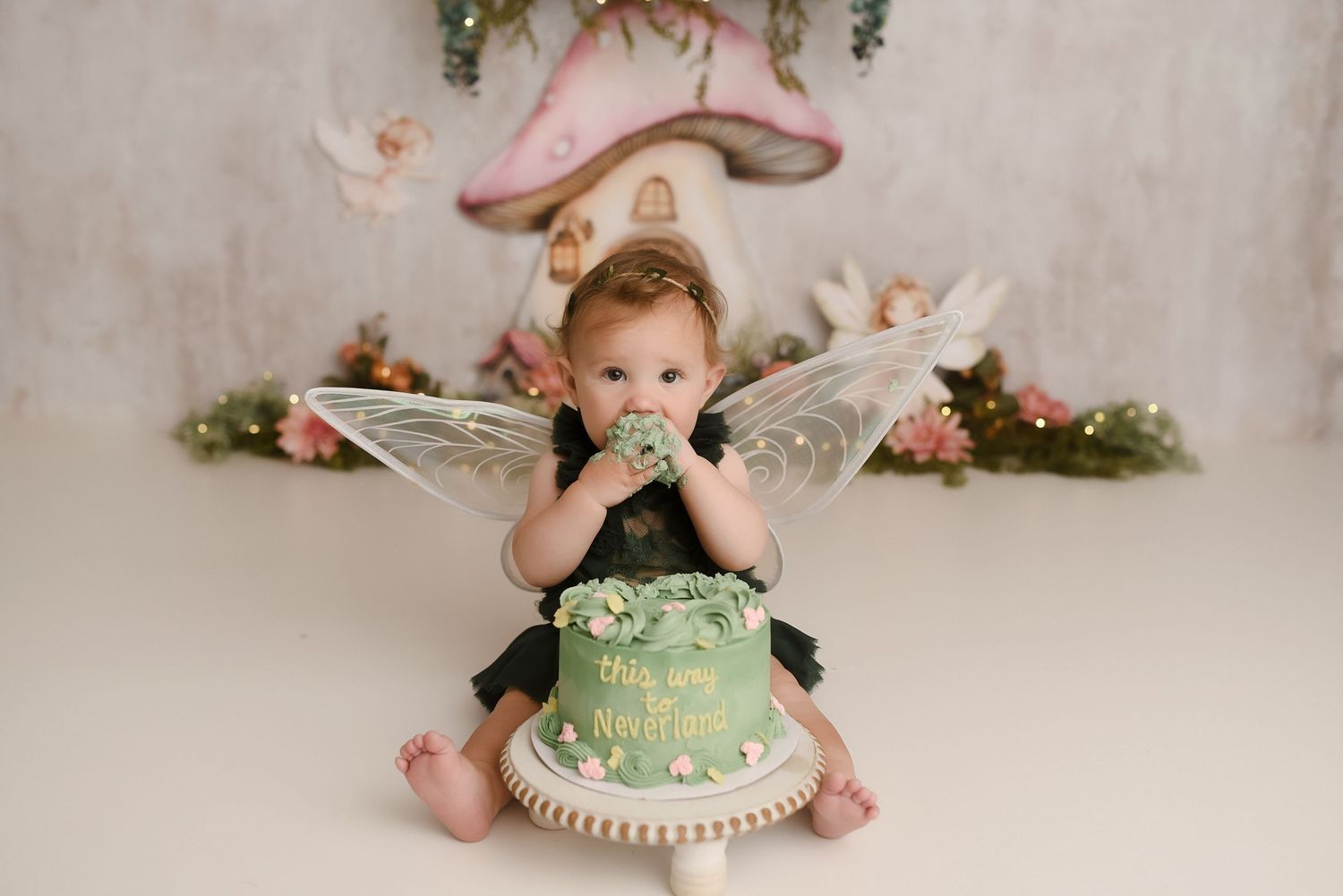 Baby girl wearing fairy wings eating green first birthday cake in enchanted mushroom garden photo session.