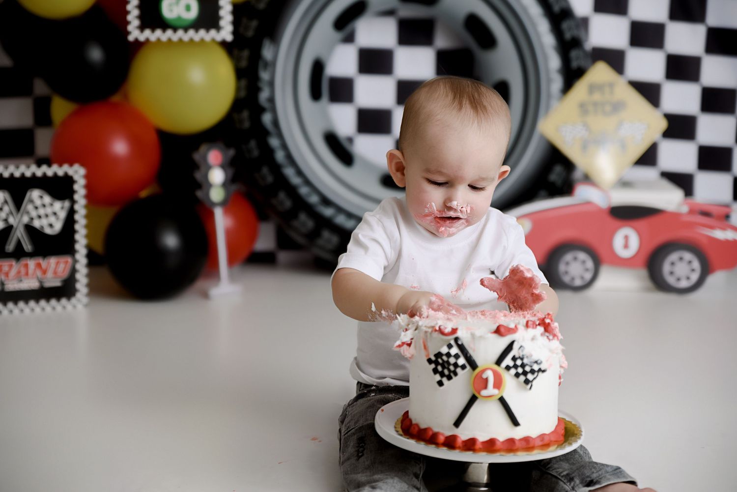 Baby boy smashing race car themed first birthday cake with checkered flags and balloons.