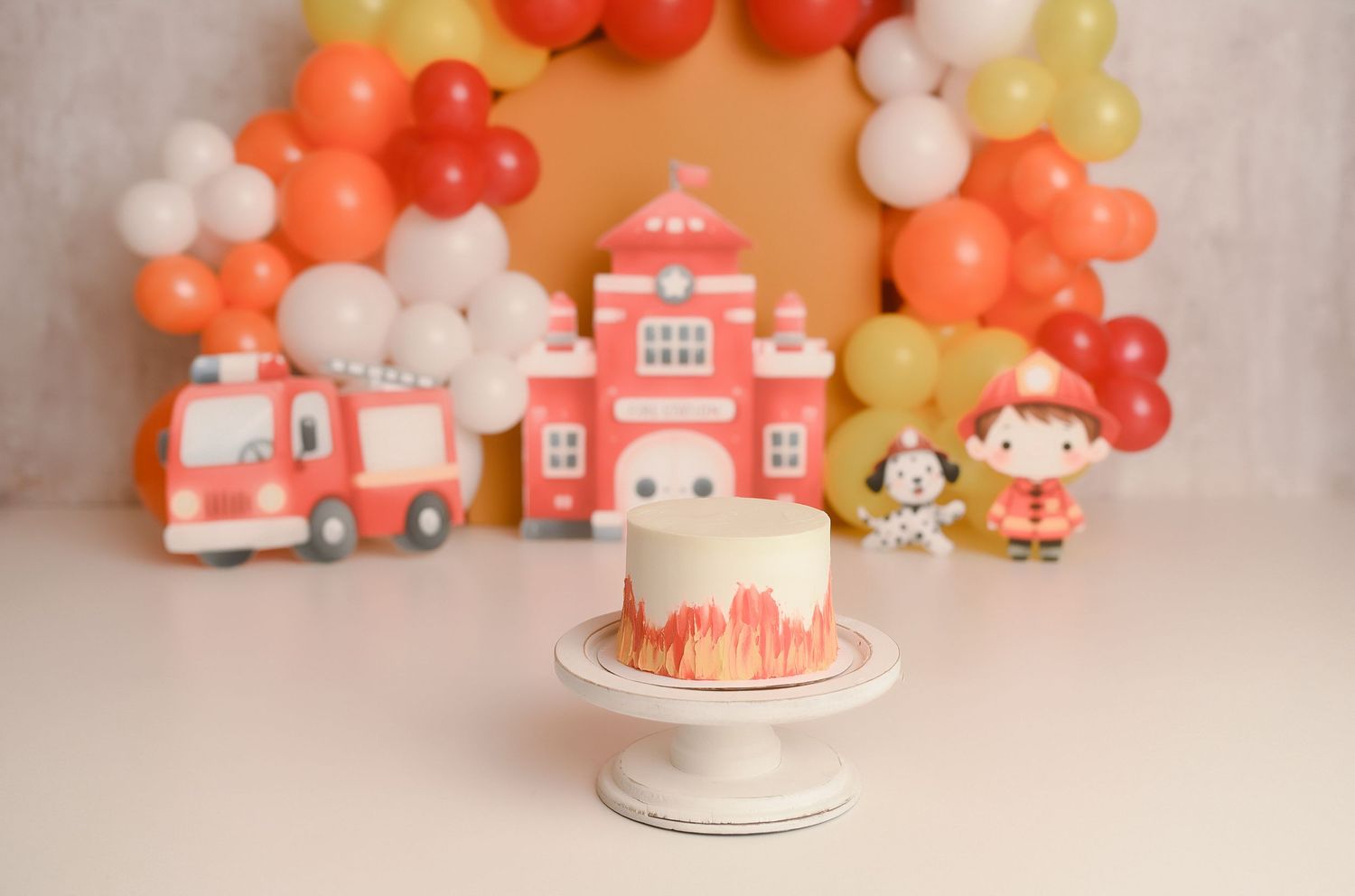 Fire station themed first birthday cake smash setup with balloon arch, fire truck props, and white fondant cake.