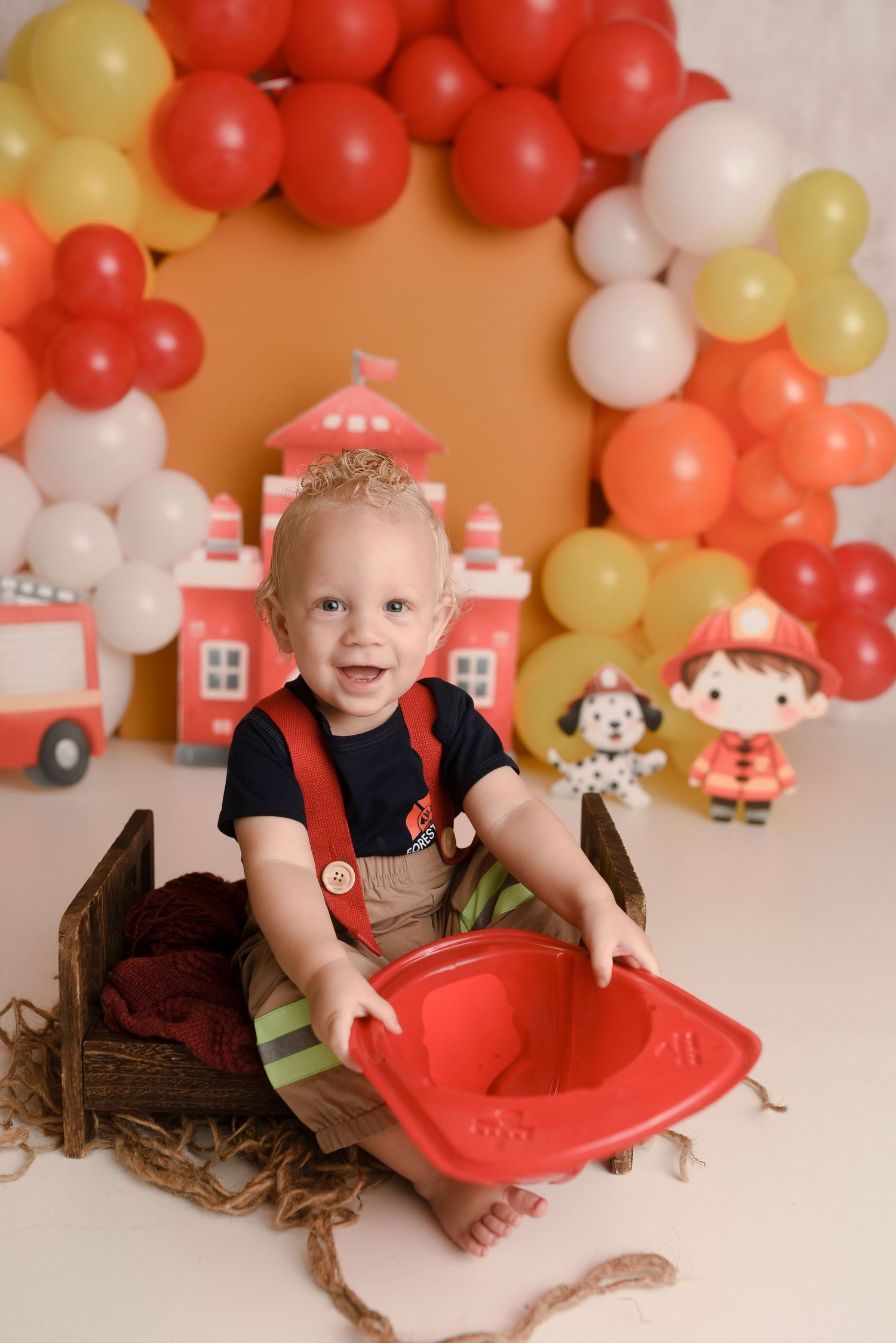 Smiling baby boy in firefighter costume sits in wooden crate with red helmet, surrounded by colorful balloon arch backdrop.