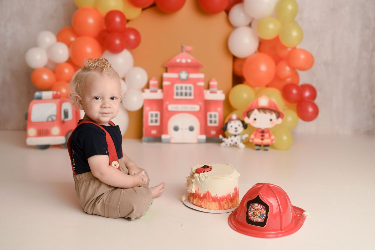 Baby boy in firefighter-themed first birthday cake smash session with fire truck and balloon garland backdrop.