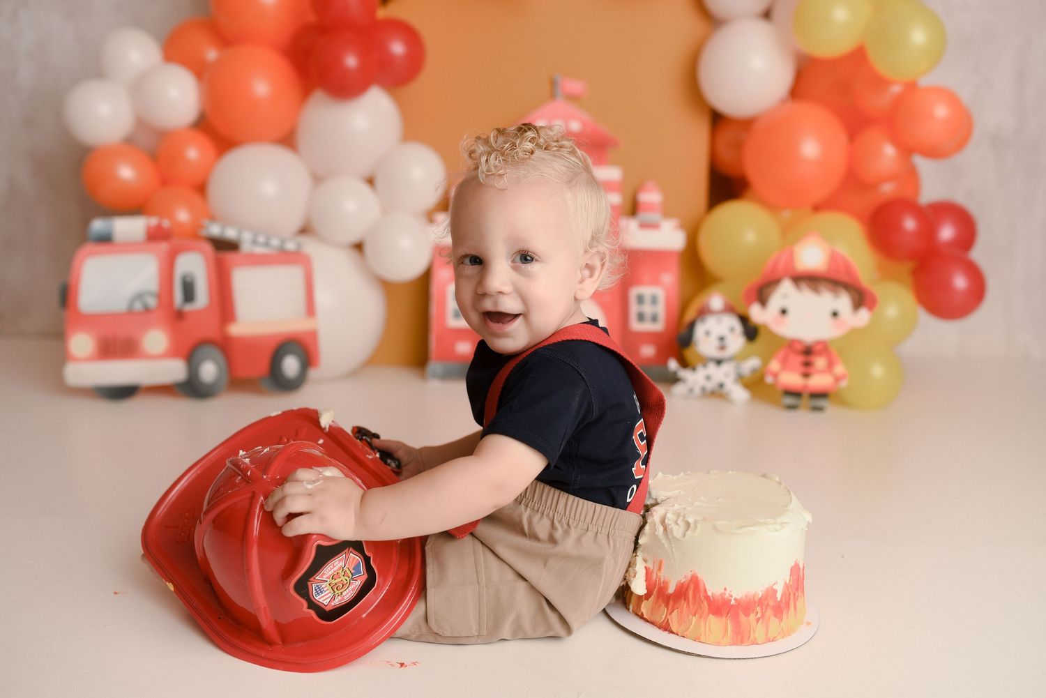 Toddler boy holding red firefighter helmet during cake smash photo session with fire truck balloon backdrop.