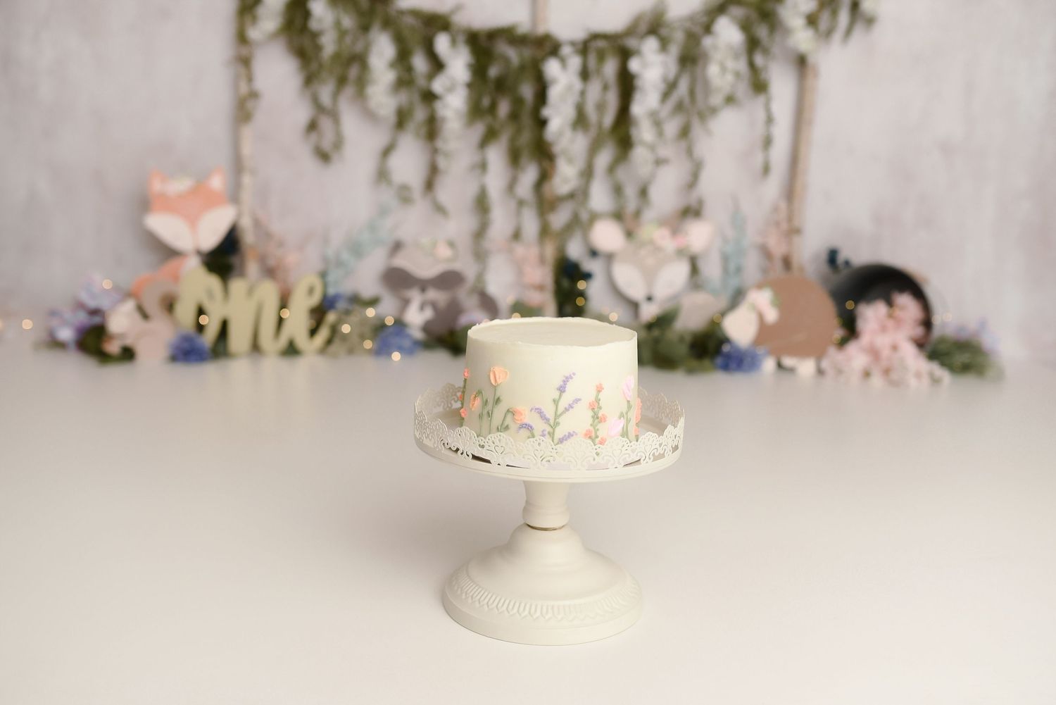 Elegant white first birthday cake with floral details on a pedestal stand with woodland animal party backdrop.