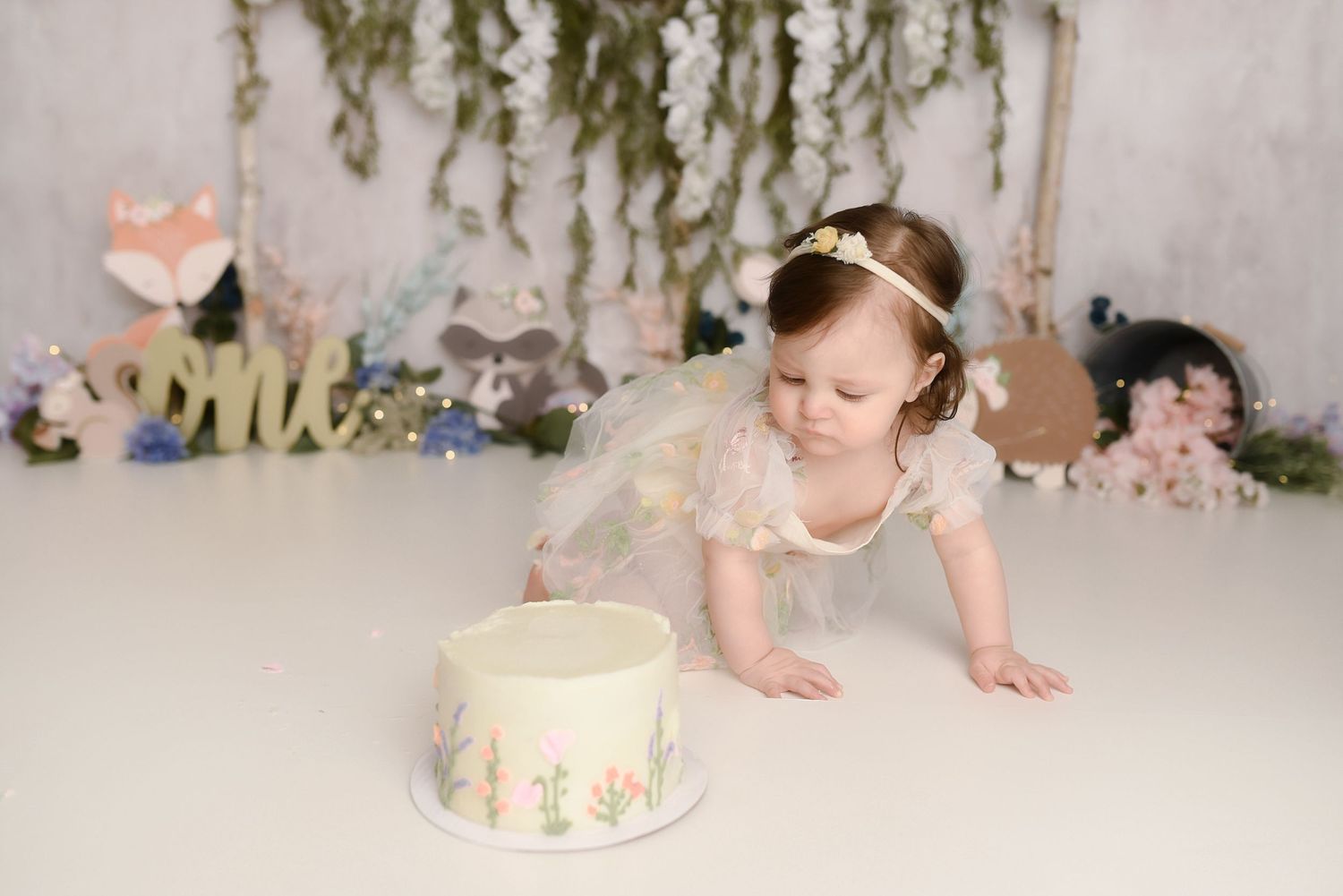 Baby girl in white dress reaching toward decorated first birthday cake at woodland themed cake smash session.