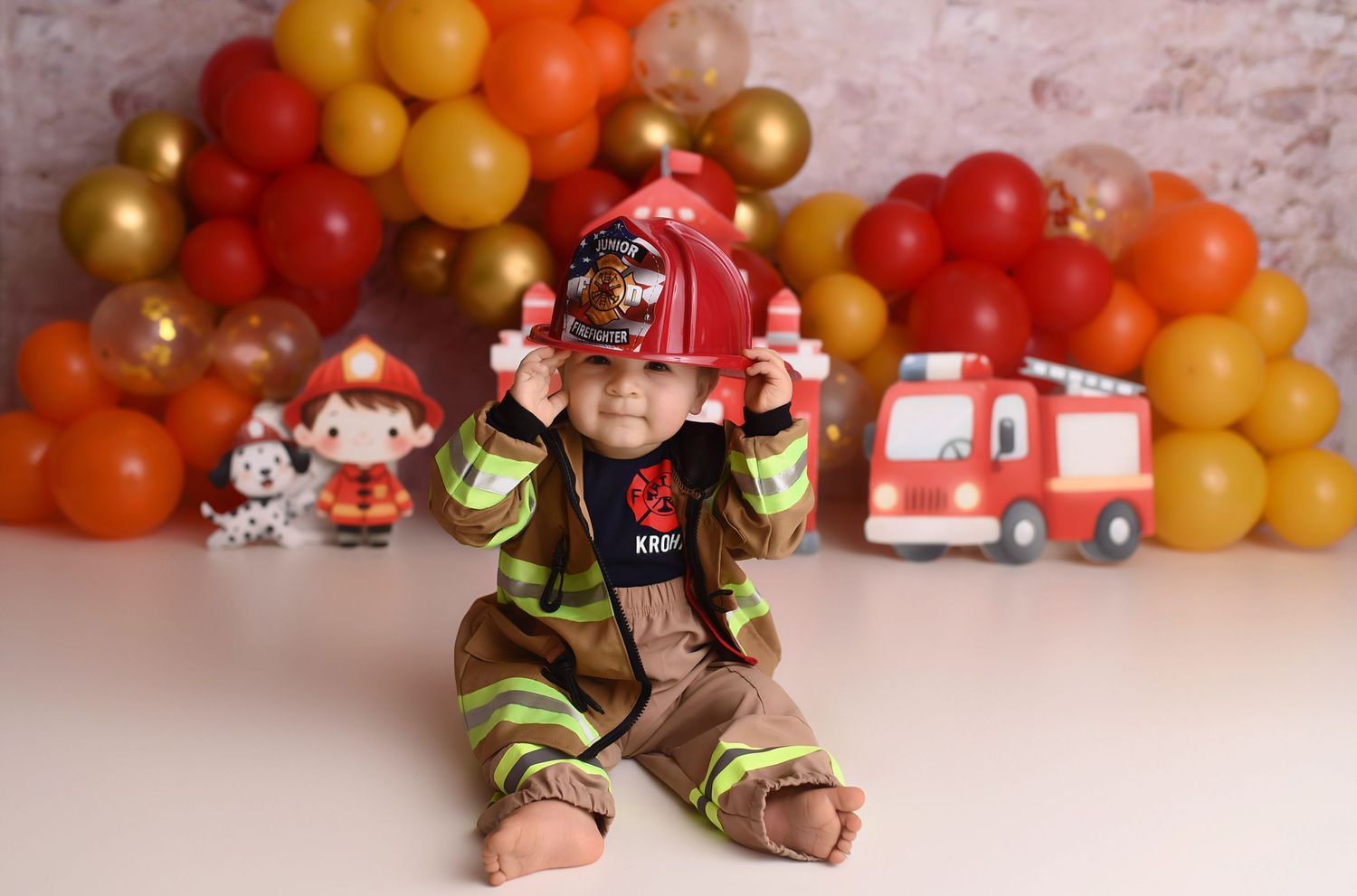 Cute baby boy in firefighter costume holds red helmet at fire truck themed first birthday session.