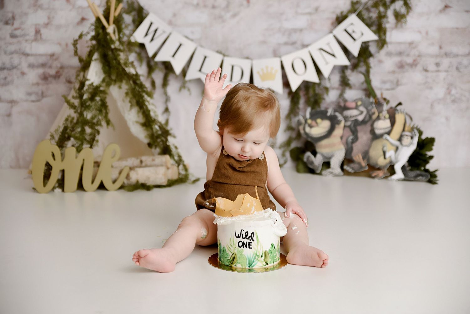 Baby smashing Wild One cake with teepee and jungle animals banner backdrop.