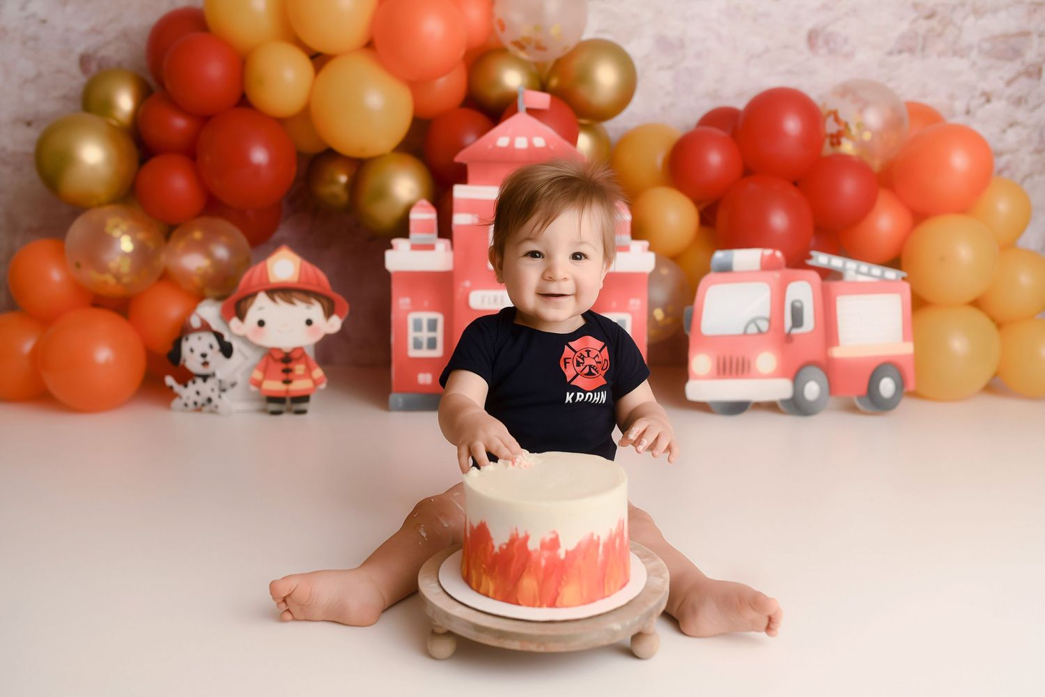 Baby boy in navy onesie reaches for red and white smash cake at fire truck birthday photo session.