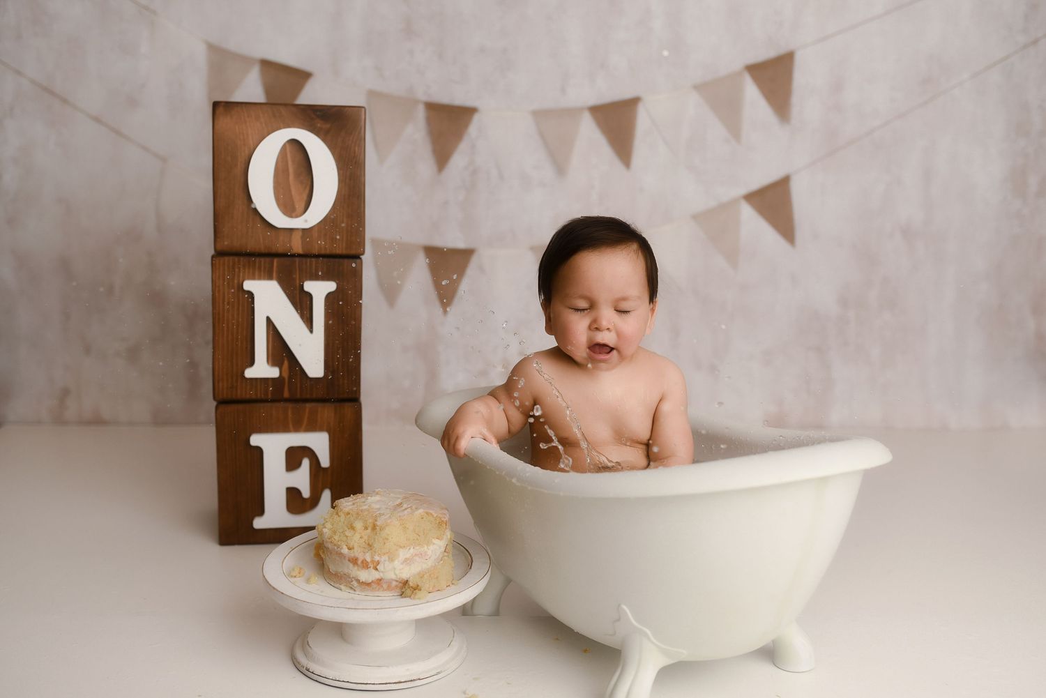 Baby in a white mini bathtub beside stacked ONE blocks and bunting for a first birthday cake smash photo.