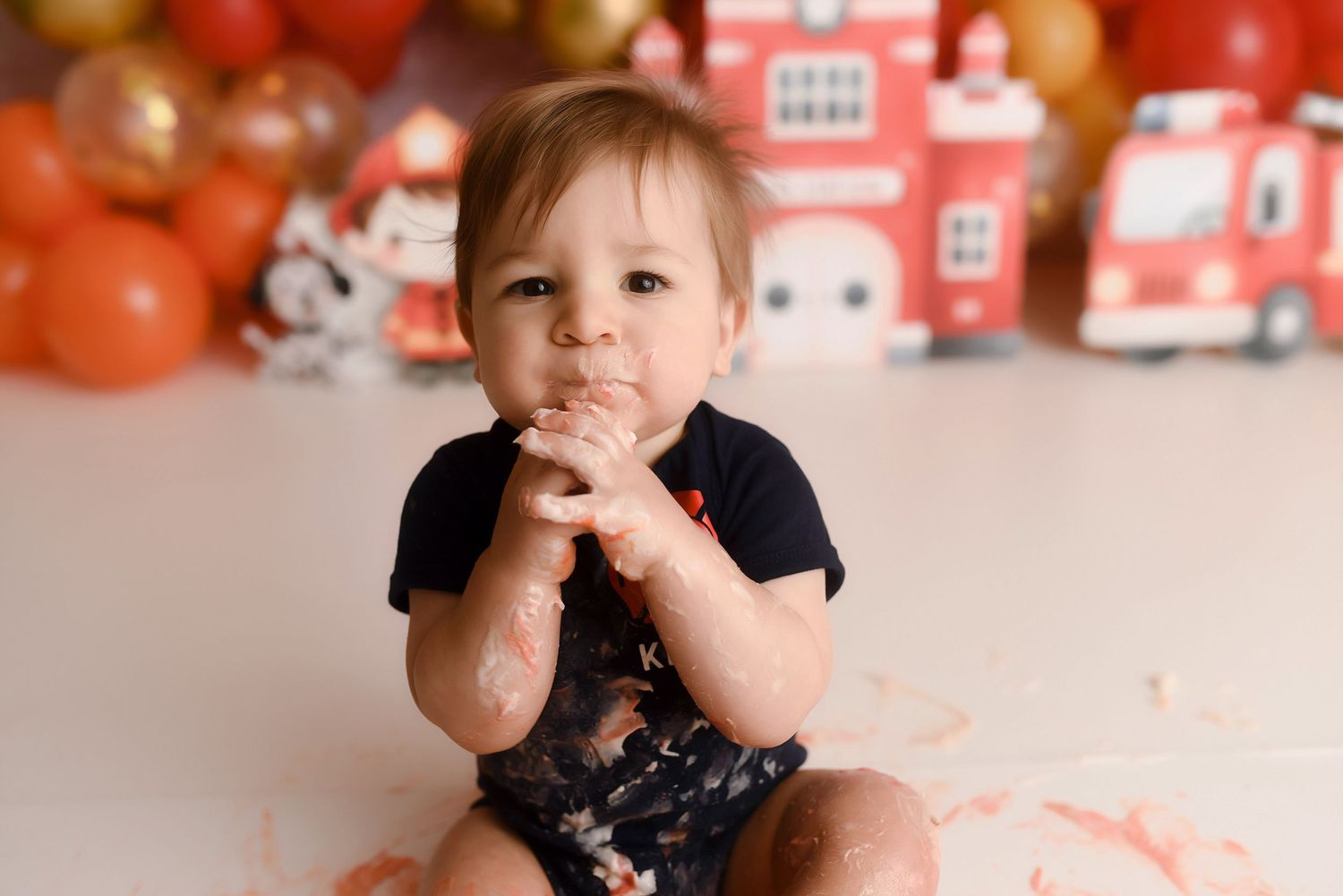 Messy baby boy eats cake with hands during fire truck themed first birthday cake smash photography.