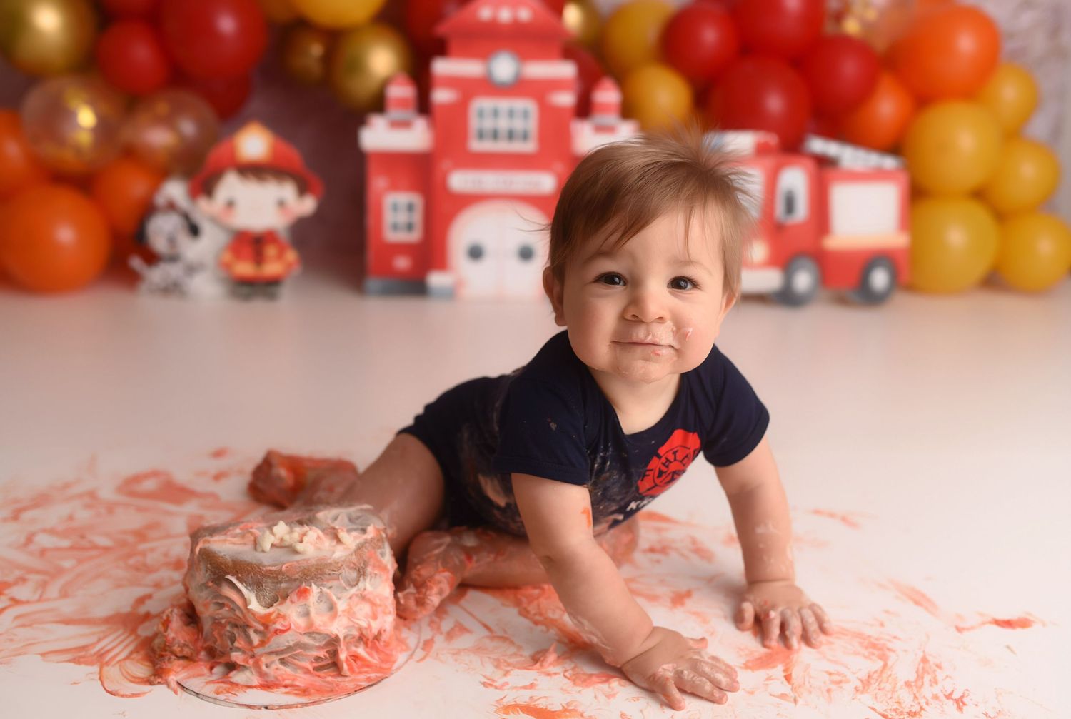 Baby boy crawls through smashed birthday cake at colorful fire truck themed cake smash photo session.