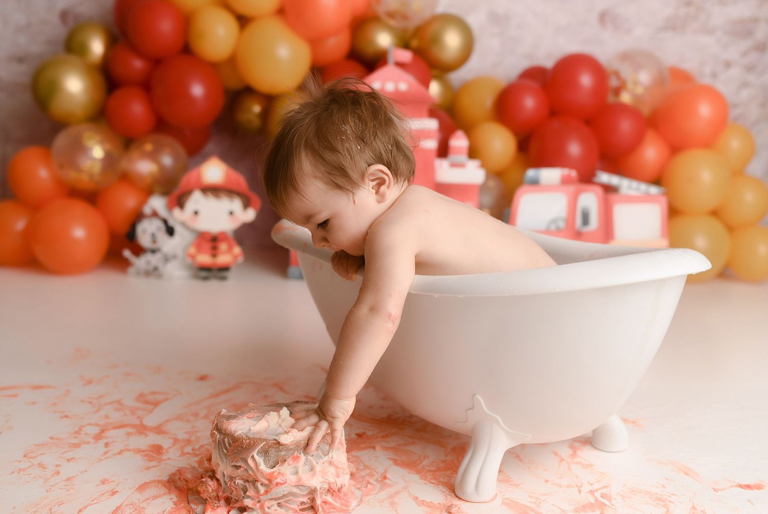 Baby sits in white clawfoot tub surrounded by cake smash mess at fire truck themed photo session.