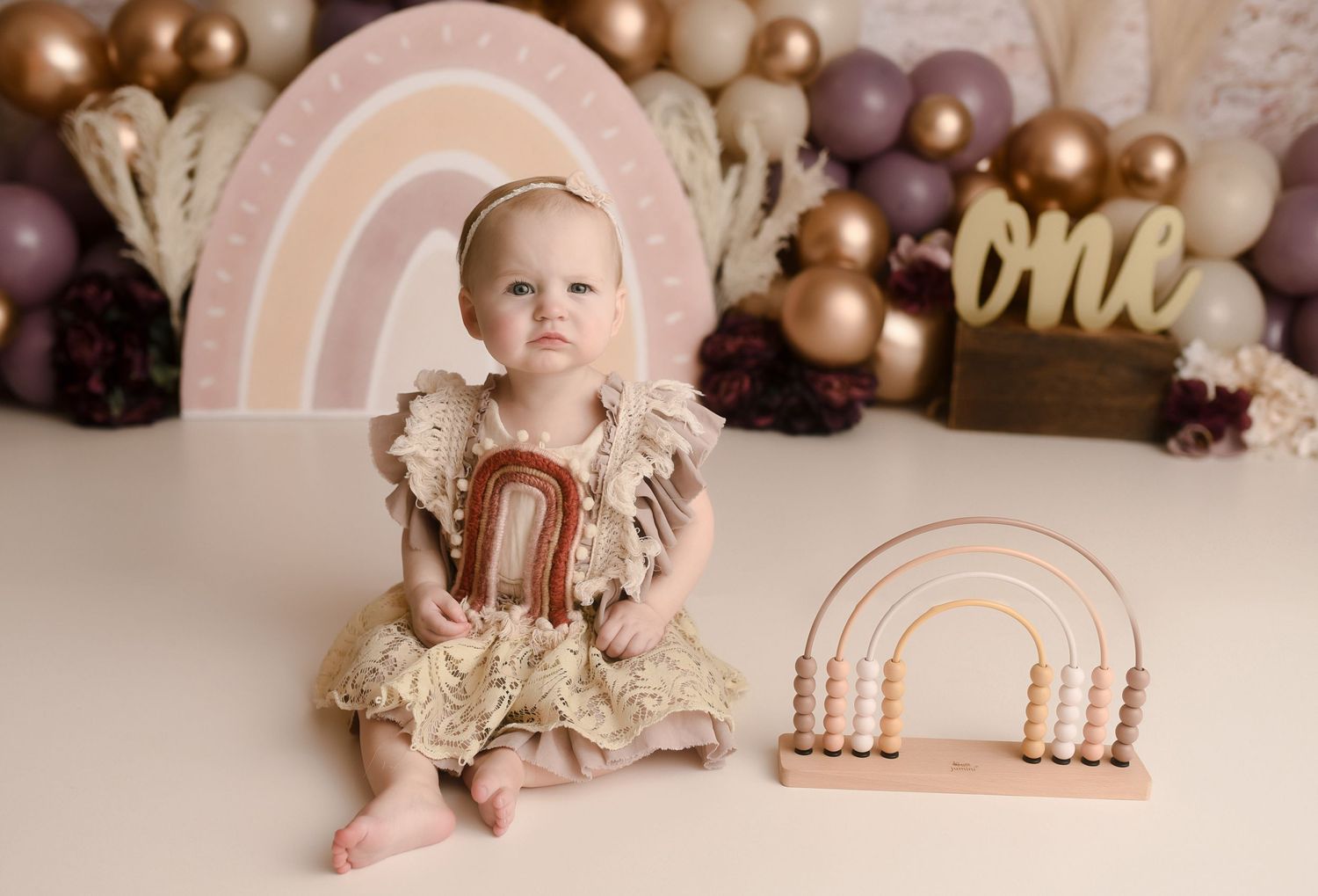 Baby girl in gold lace romper sitting with rainbow toy and boho balloon backdrop.
