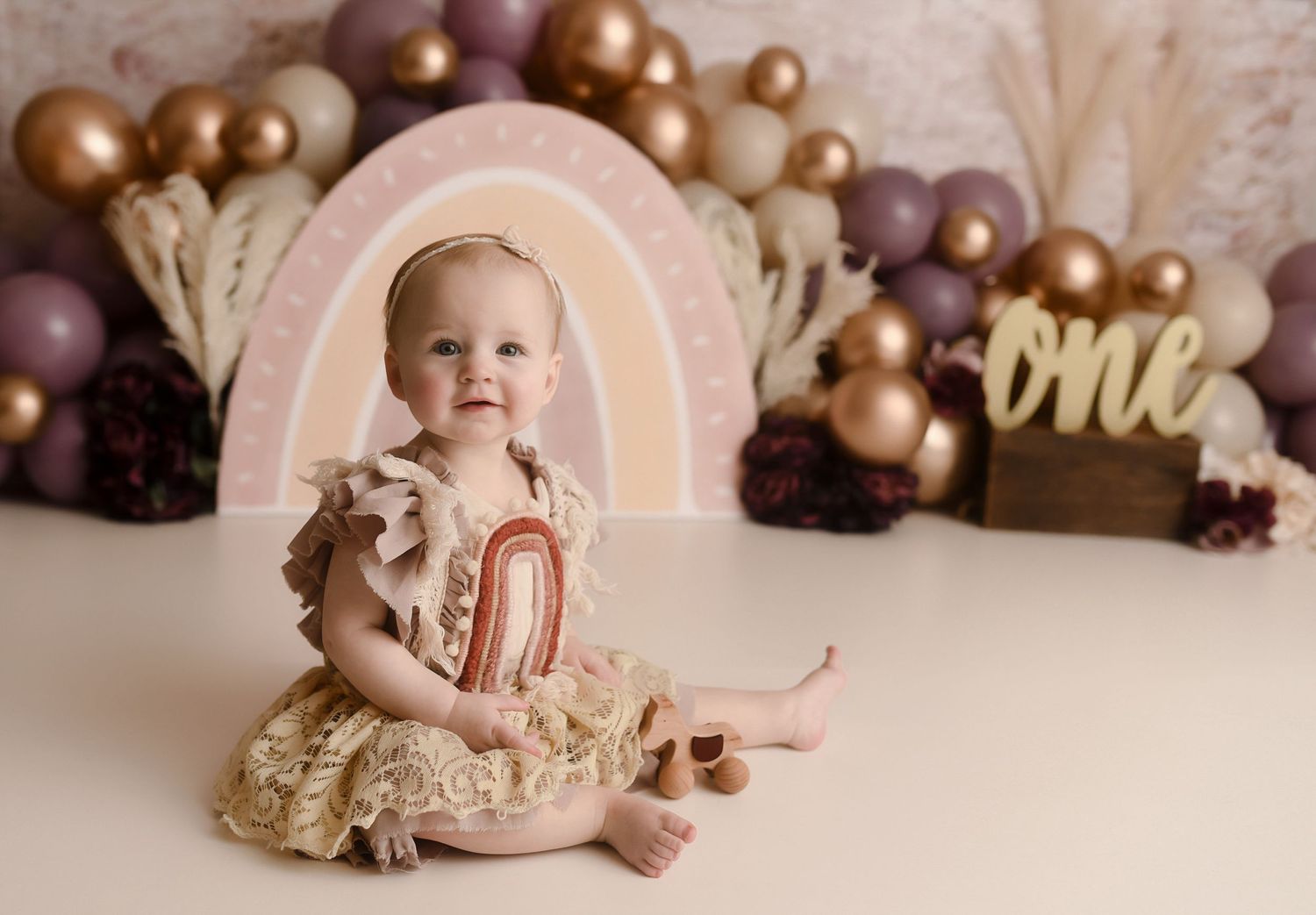 Smiling baby girl in gold lace romper holding rainbow toy with boho balloon backdrop.