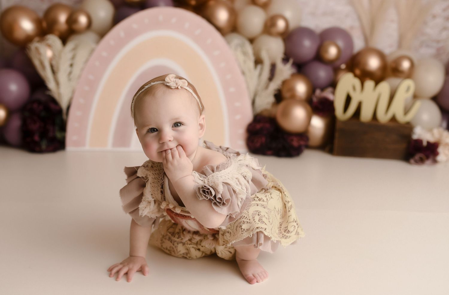 Adorable baby girl in lace romper poses at boho rainbow first birthday cake smash photo session.